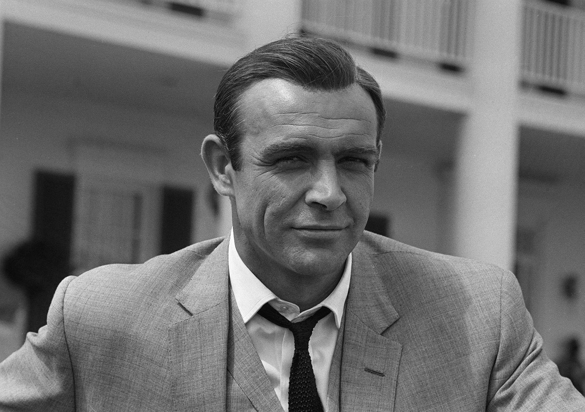 Sean Connery 桌布 壁紙 — 一張清晰的黑白肖恩·康納利肖像，身穿西裝，帶著微妙的笑意，背景是模糊的建築物。