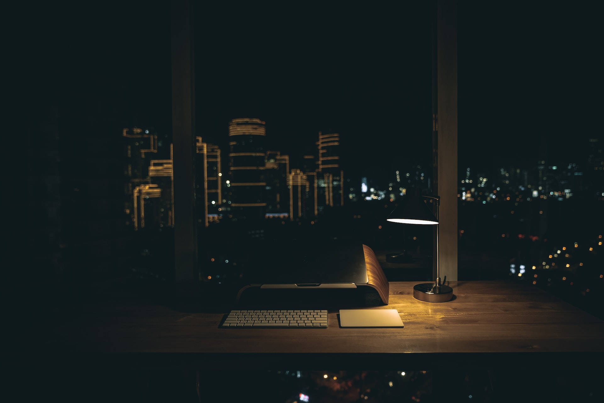 辦公室 桌布 壁紙 — 一張溫暖照明的書桌，放著筆記型電腦和燈，透過大窗戶俯瞰夜晚閃爍的廣闊城市景觀。