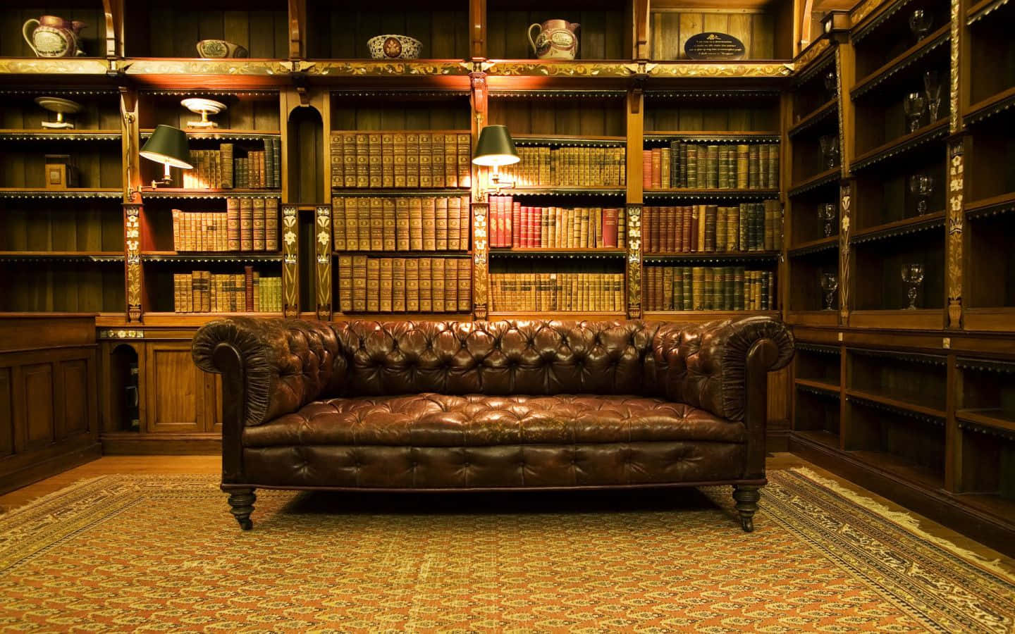 Library 桌布 壁紙 — 一張濃郁的棕色皮革切斯特菲爾德沙發居中放在圖案地毯上，前面是高聳的木書架，裝滿了復古書籍和裝飾品。