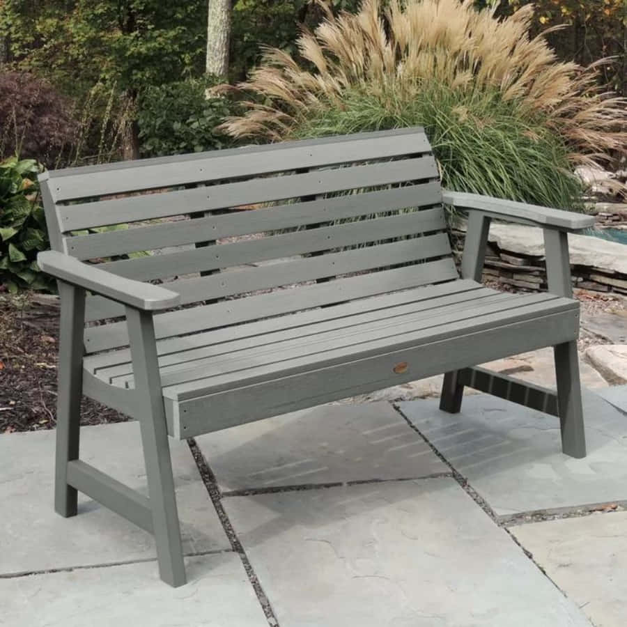 Bench 桌布 壁紙 — 一張灰色板條花園長椅放在石板上，旁邊是高高的羽毛狀草和茂密的灌木，在寧靜的後院中。