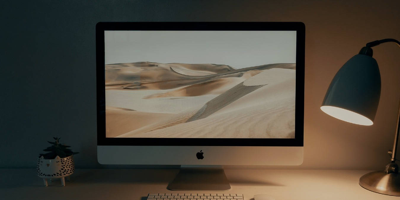 Work 桌布 壁紙 — 一張燈光昏暗的桌子上有一部iMac，顯示著寧靜的沙漠景觀，而一盞溫暖的桌燈將柔和的光線投射在一棵小盆栽上。
