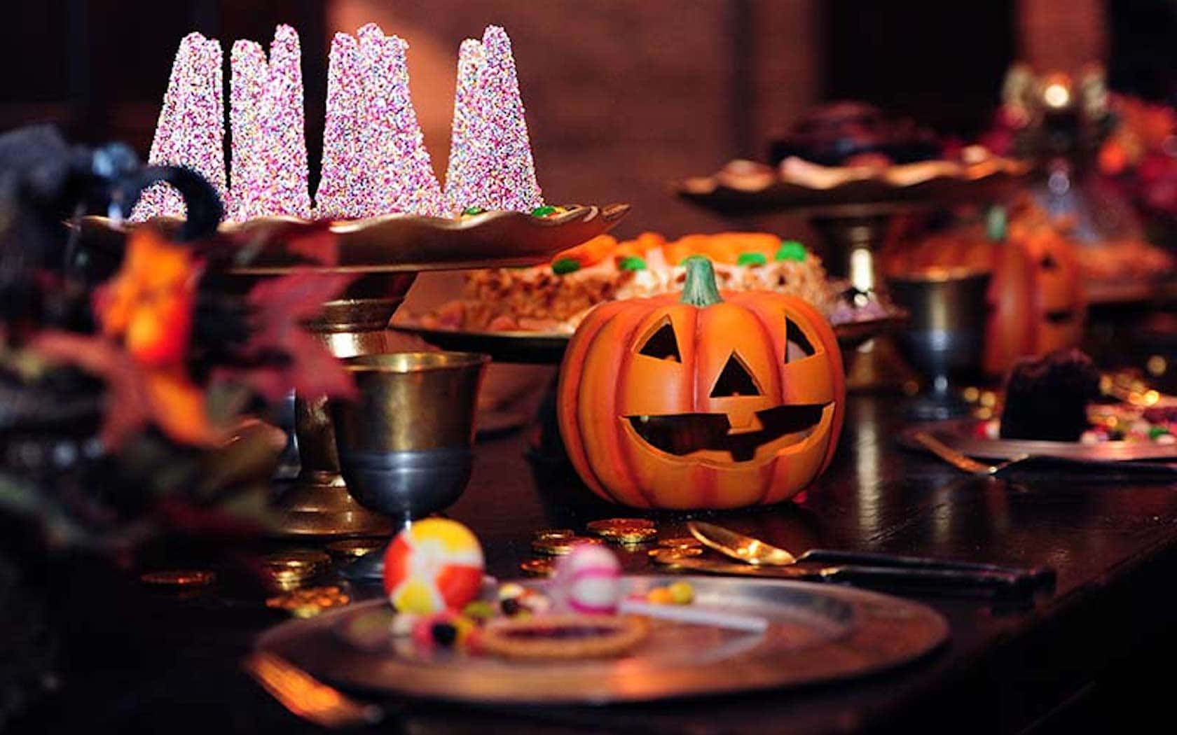 Harry Potter Halloween 桌布 壁紙 — 一張燈光昏暗的萬聖節餐桌，以雕刻南瓜為中心裝飾，並在溫暖的環境燈光下擺放閃閃發光的錐形糖果。