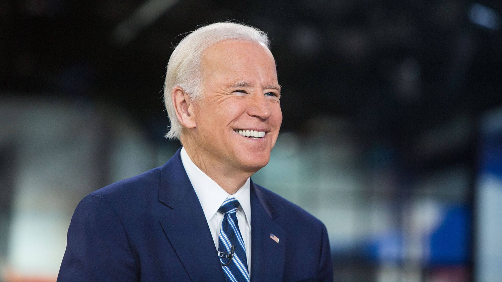 Biden 桌布 壁紙 — 一張特寫照片，一位微笑的老年男士身穿深色西裝和條紋領帶，背景是模糊的室內環境。
