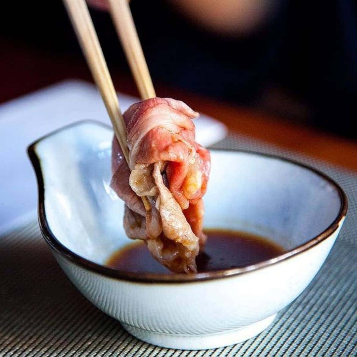 肉 桌布 壁紙 — 一張特寫照片，筷子從裝滿深色醬汁的陶瓷蘸醬碗中夾起薄切生牛肉。