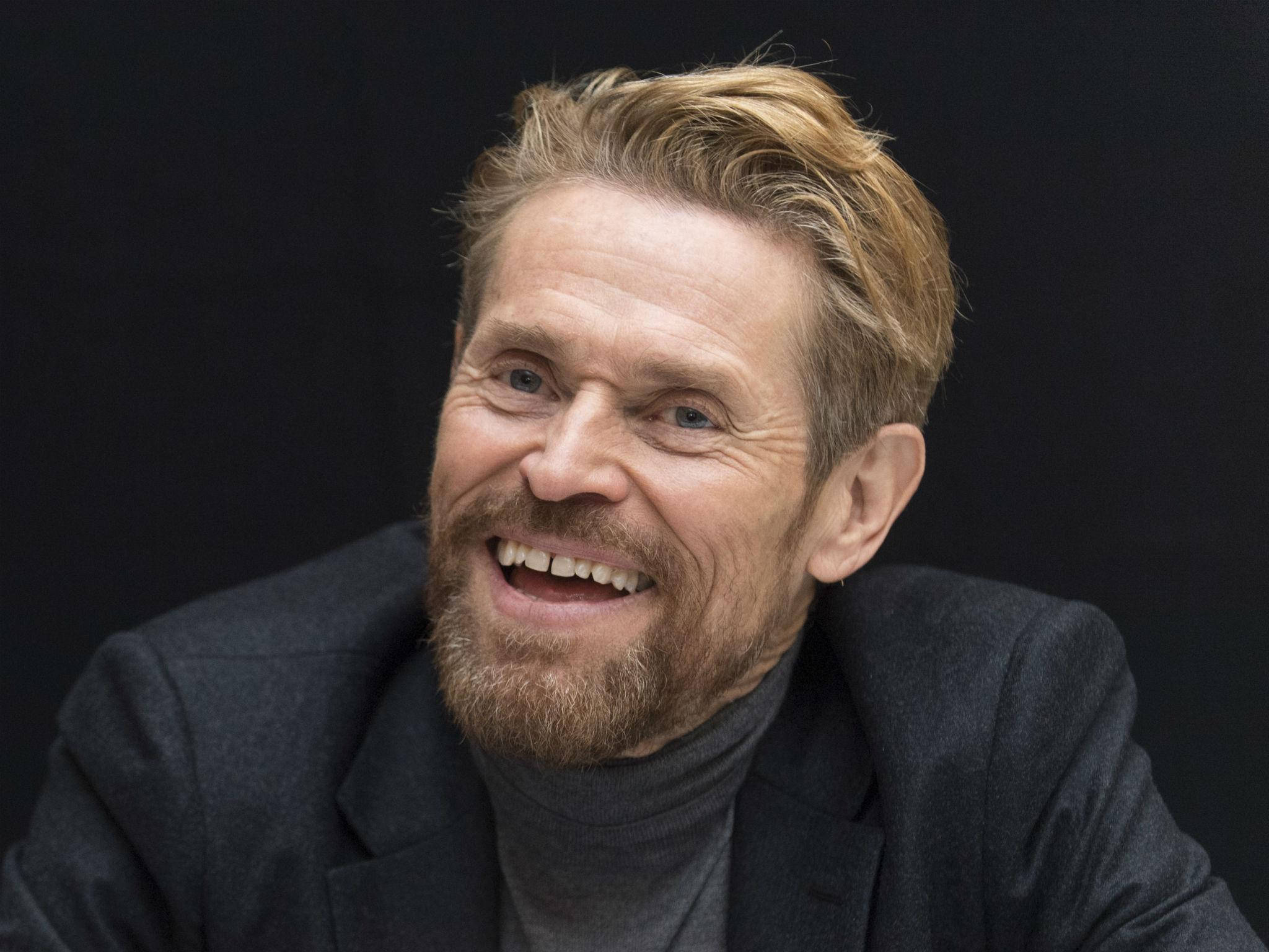 Willem Dafoe 桌布 壁紙 — 一張特寫照片，顯示一位留著鬍子、金髮的男子在深色背景前開懷大笑。
