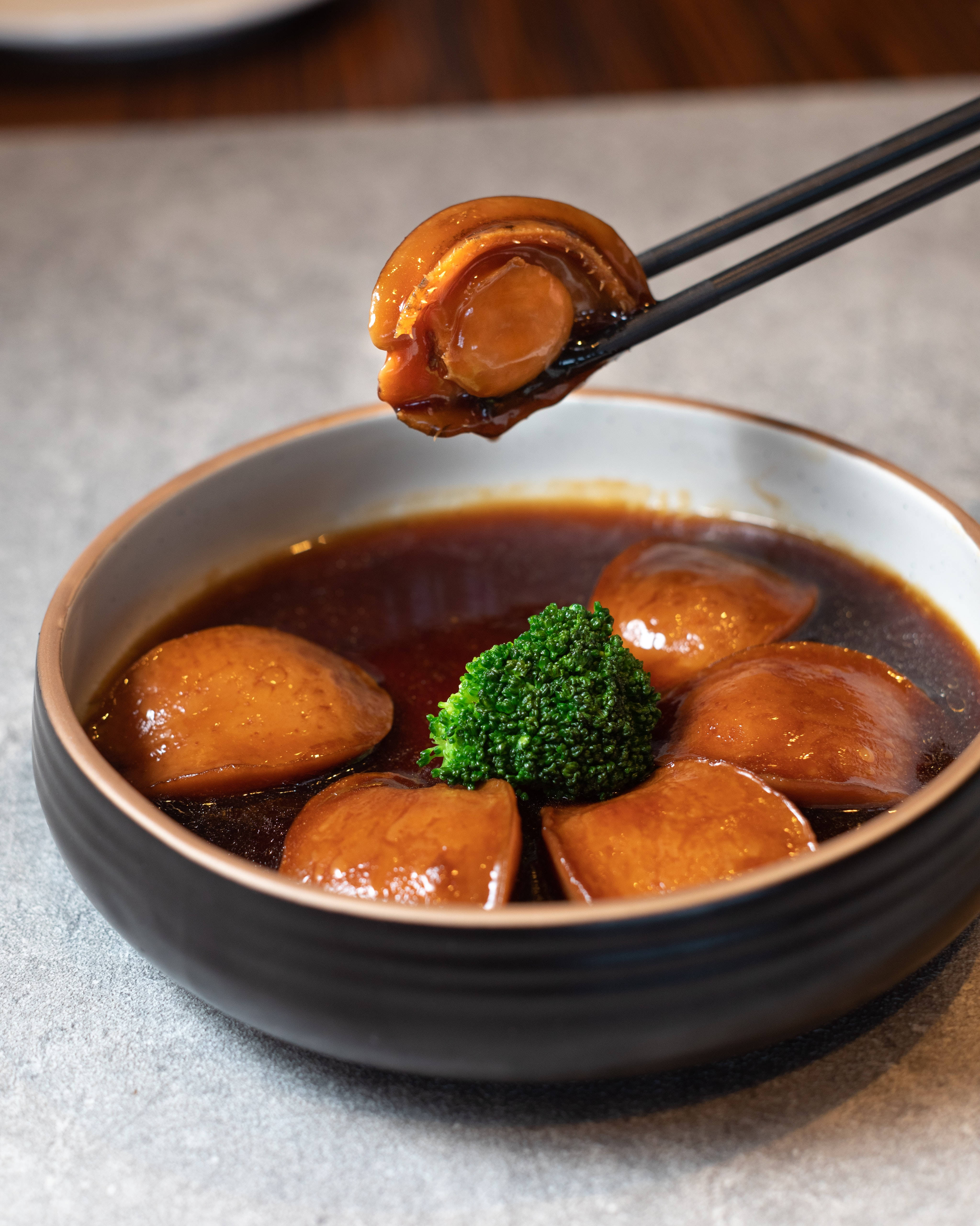 Food Photography 桌布 壁紙 — 一張特寫照片，顯示用筷子夾起光亮的燜鮑魚，放在深色湯碗上方，碗邊點綴著鮮綠的西蘭花。
