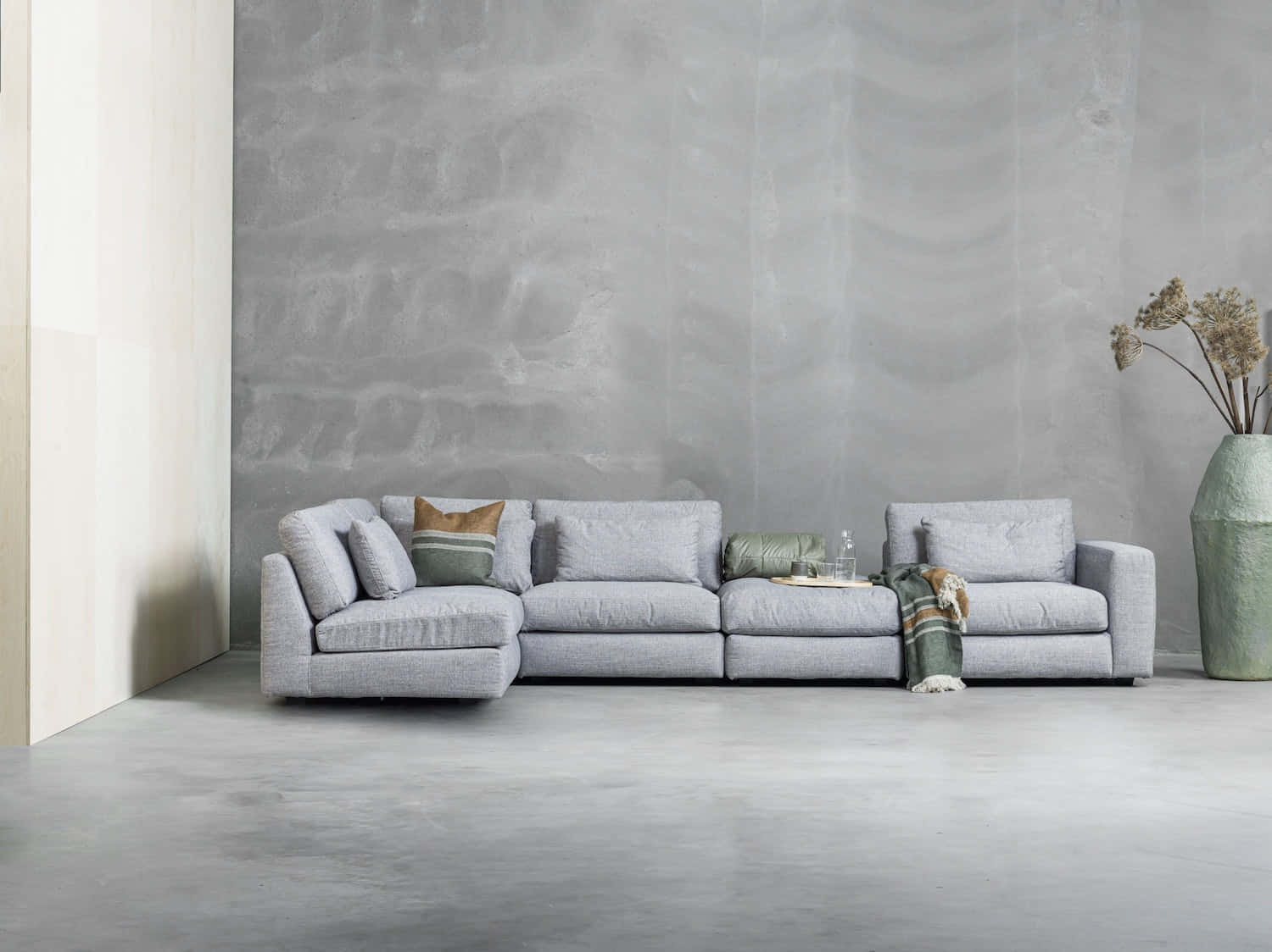 Living Room 桌布 壁紙 — 一張現代淺灰色組合沙發靠著紋理混凝土牆，在一個極簡主義的客廳中，配有一個高大的綠色花瓶，裡面插著乾花。