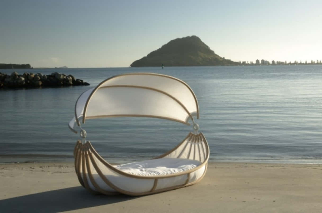 Beach Daybed 桌布 壁紙 — 一張現代白色吊床椅（附有頂篷）放置在沙灘上，面向平靜的海水，遠處有一座綠色島嶼，天空清澈湛藍。