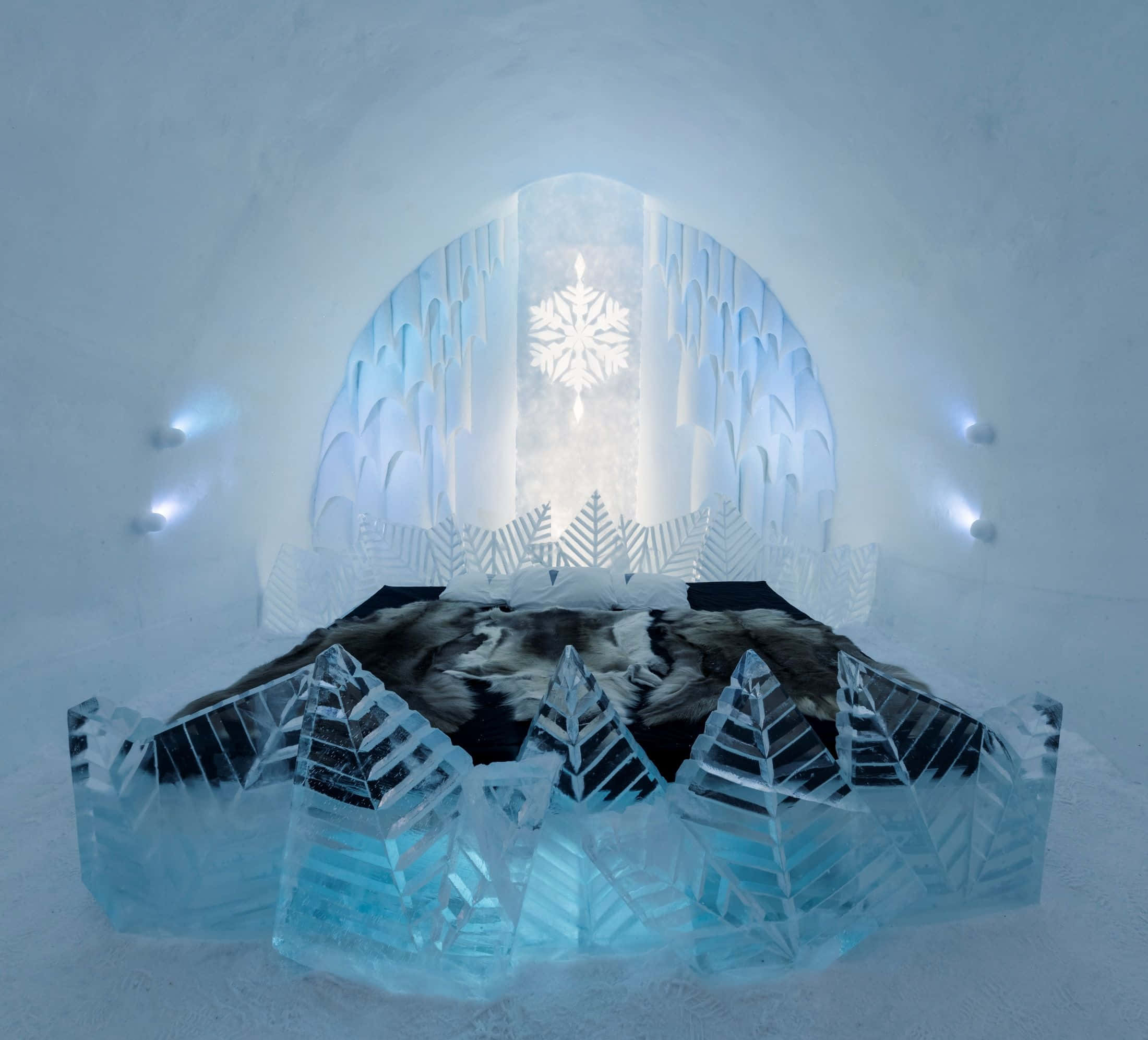Ice Hotel 桌布 壁紙 — 一張由雕刻冰晶框住的床，位於一個發光的冰拱門下，拱門上有雪花圖案，在一個冰冷的冰屋房間裡。