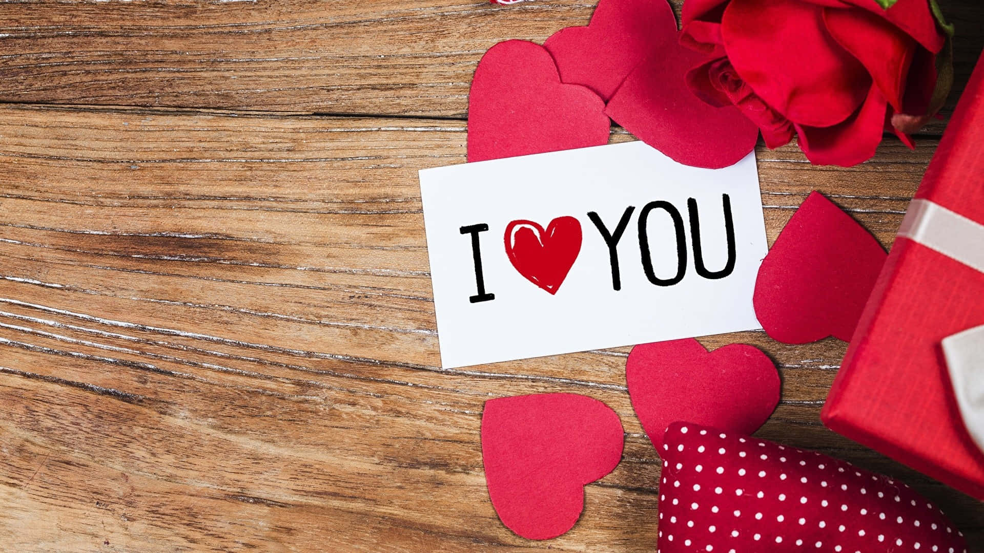 我愛你 桌布 壁紙 — 一張白色卡片，上面寫著 I heart YOU，放在鄉村風木頭上，周圍環繞著紅色紙心、玫瑰和禮物盒。