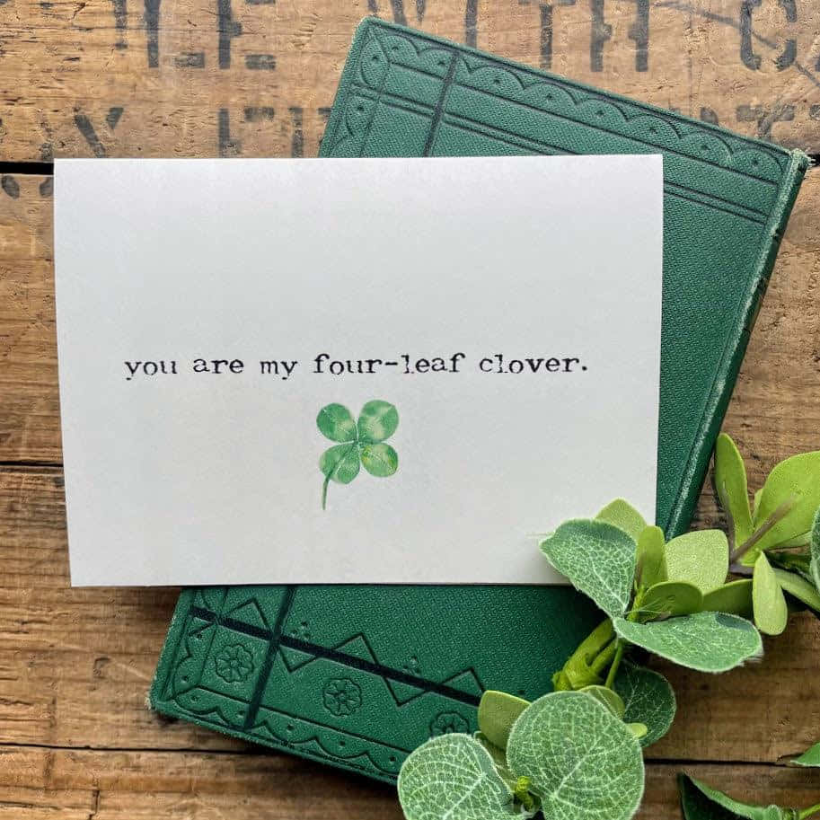 Four Leaf Clover 桌布 壁紙 — 一張白色卡片，上面有四葉草和文字，放在一本綠色書上，書放在質樸的木頭上，周圍是新鮮的綠色植物。