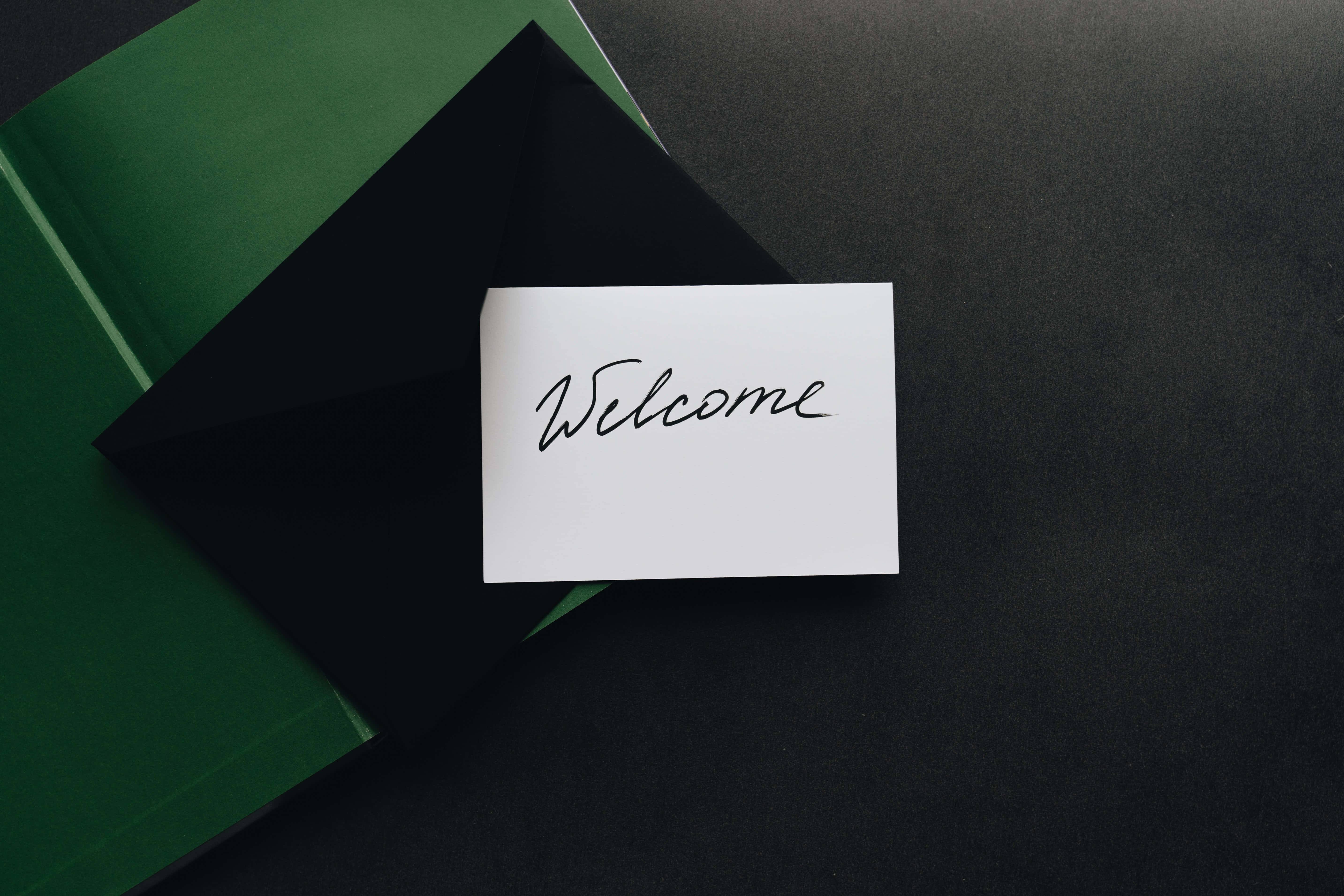 Welcome 桌布 壁紙 — 一張白色卡片，上面有手寫的歡迎文字，放在黑色信封和綠色文件夾上，置於深色表面。
