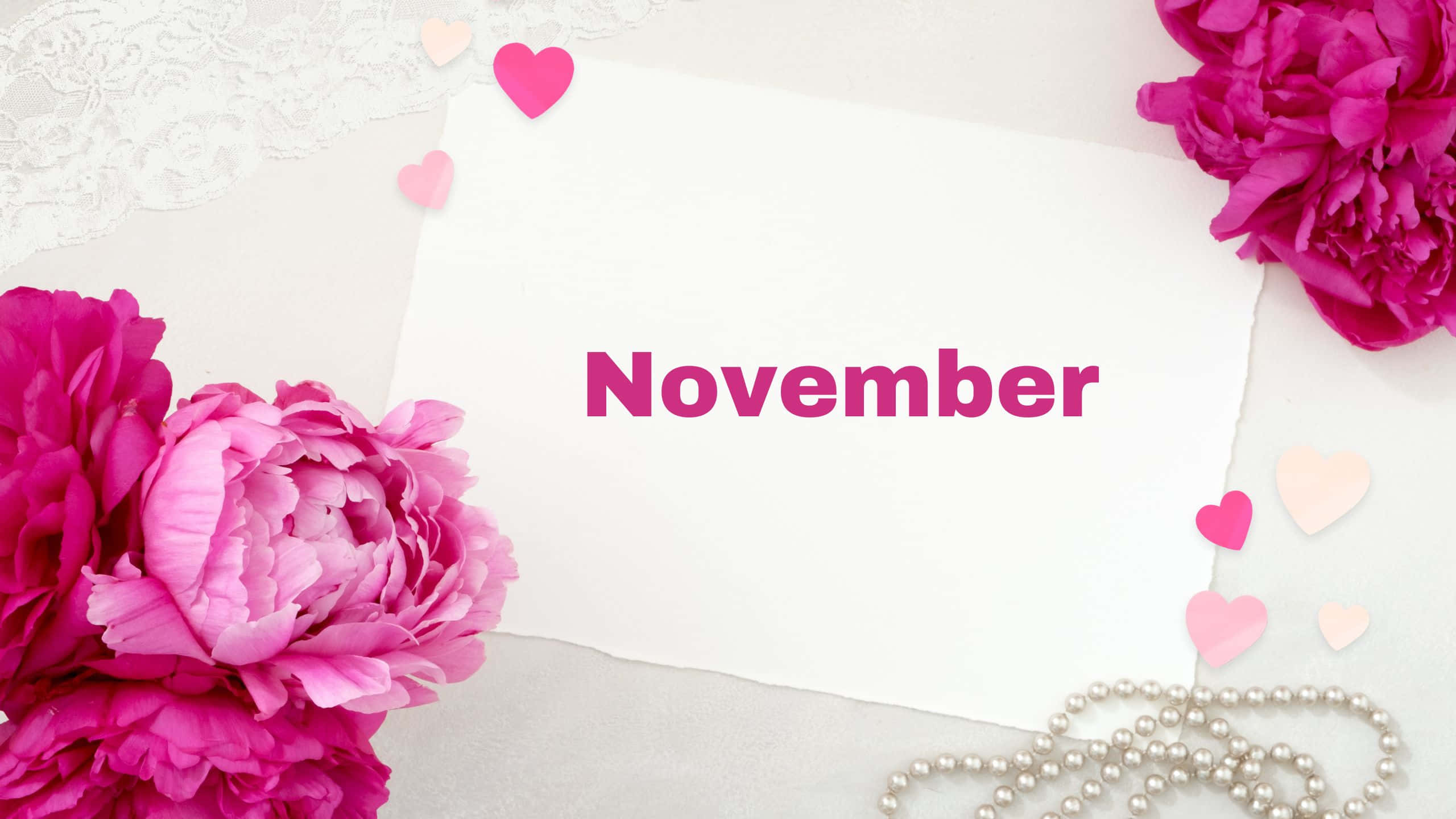 十一月 桌布 壁紙 — 一張白色卡片，上面有粉紅色的「November」文字，周圍環繞著充滿活力的粉紅色牡丹、蕾絲和珍珠珠子，放在柔軟的白色表面上。