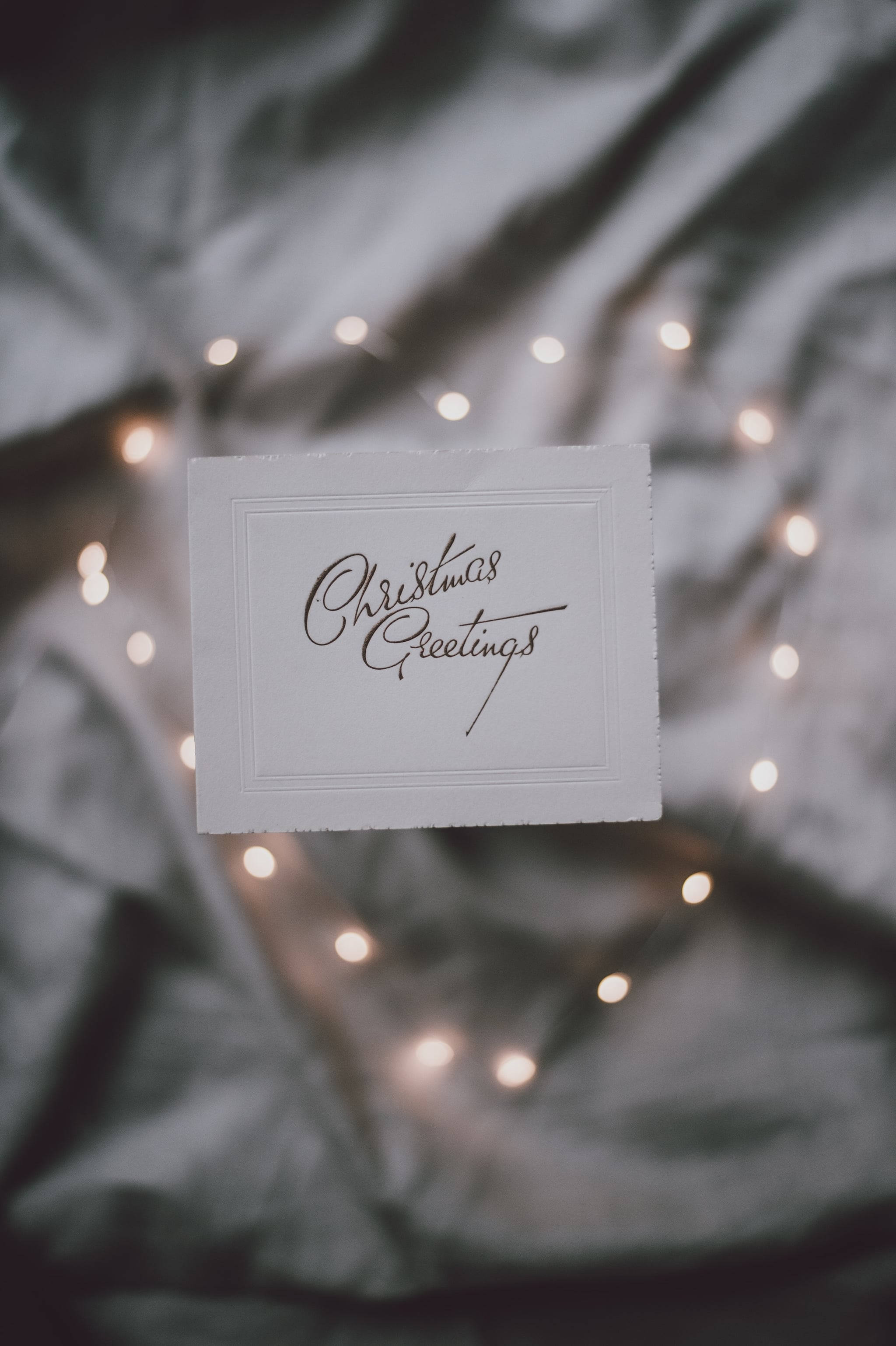 聖誕節手機 桌布 壁紙 — 一張白色聖誕卡，上面有優雅的字體寫著「Christmas Greetings」，放在柔軟的灰色布料背景上，背景點綴著閃爍的仙女燈。