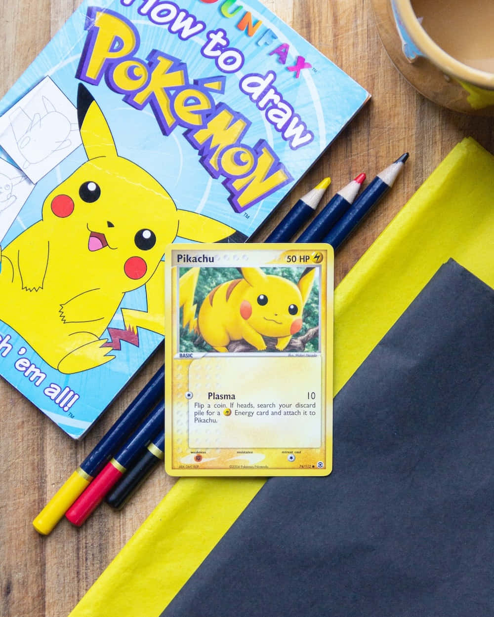 Pokemon Card 桌布 壁紙 — 一張皮卡丘交換卡和繪畫簿放在木桌上，旁邊有彩色鉛筆和一個杯子。