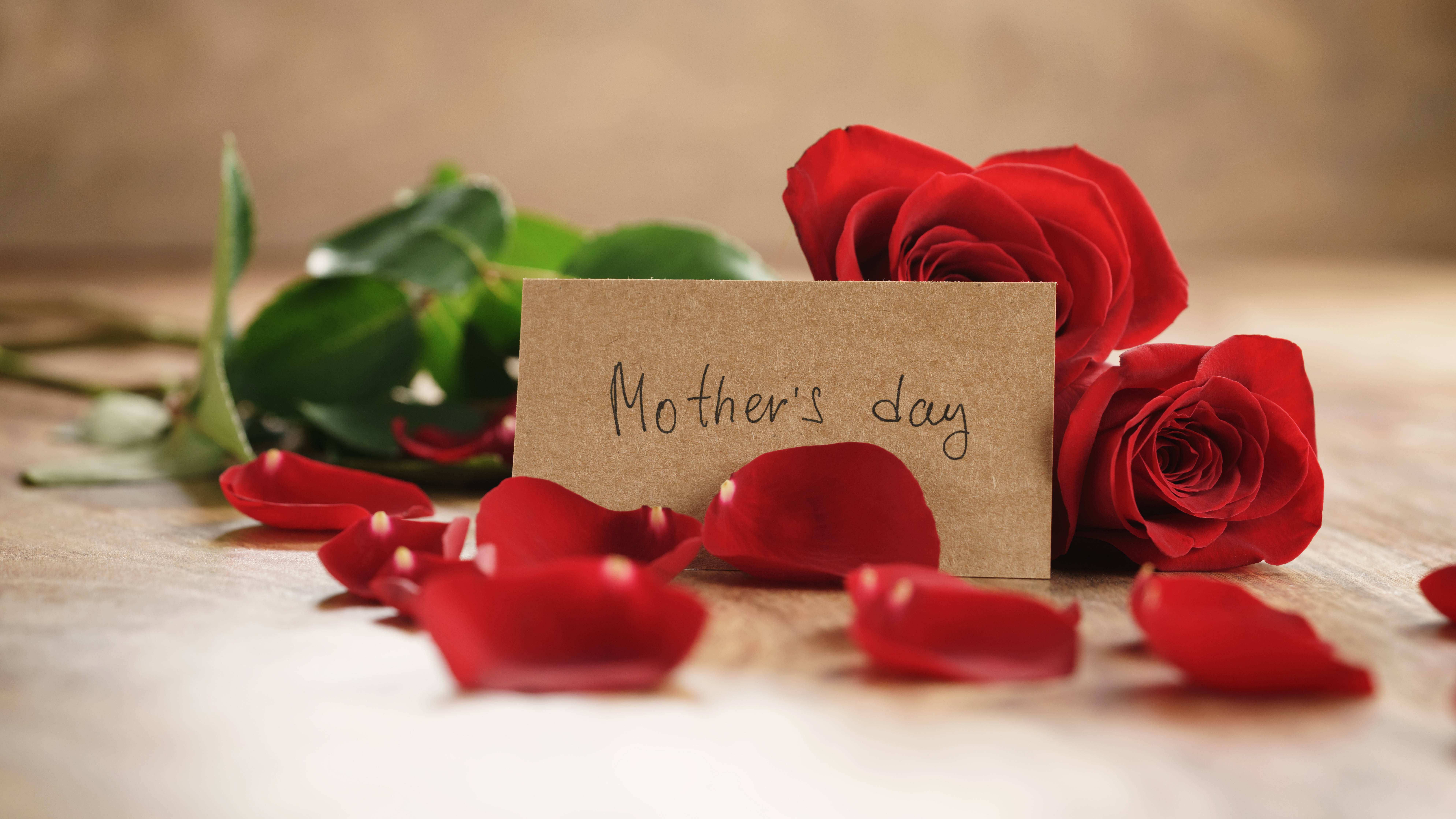 Happy Mothers Day 桌布 壁紙 — 一張窩心的母親節卡片，依偎在鮮豔的紅玫瑰和散落的花瓣之間，置於溫暖的木質表面上。
