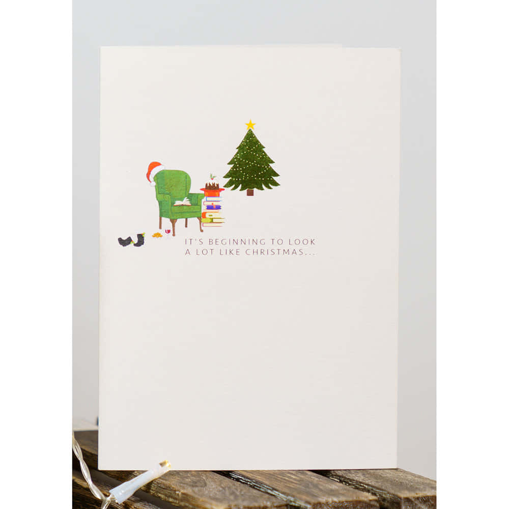 Christmas Card 桌布 壁紙 — 一張節日插圖賀卡顯示一張綠色扶手椅，上面有聖誕帽，旁邊有一棵裝飾好的聖誕樹和一疊書。