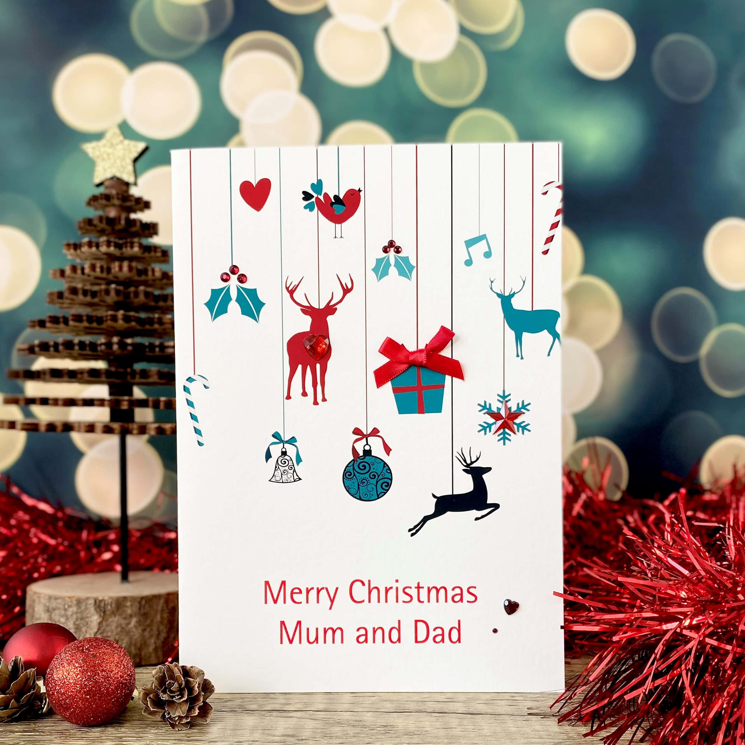 Christmas Card 桌布 壁紙 — 一張節日聖誕卡，掛有紅色和藍綠色裝飾品，包括馴鹿禮物和鳥類，背景為散景效果，配有紅色閃粉和巧克力樹。
