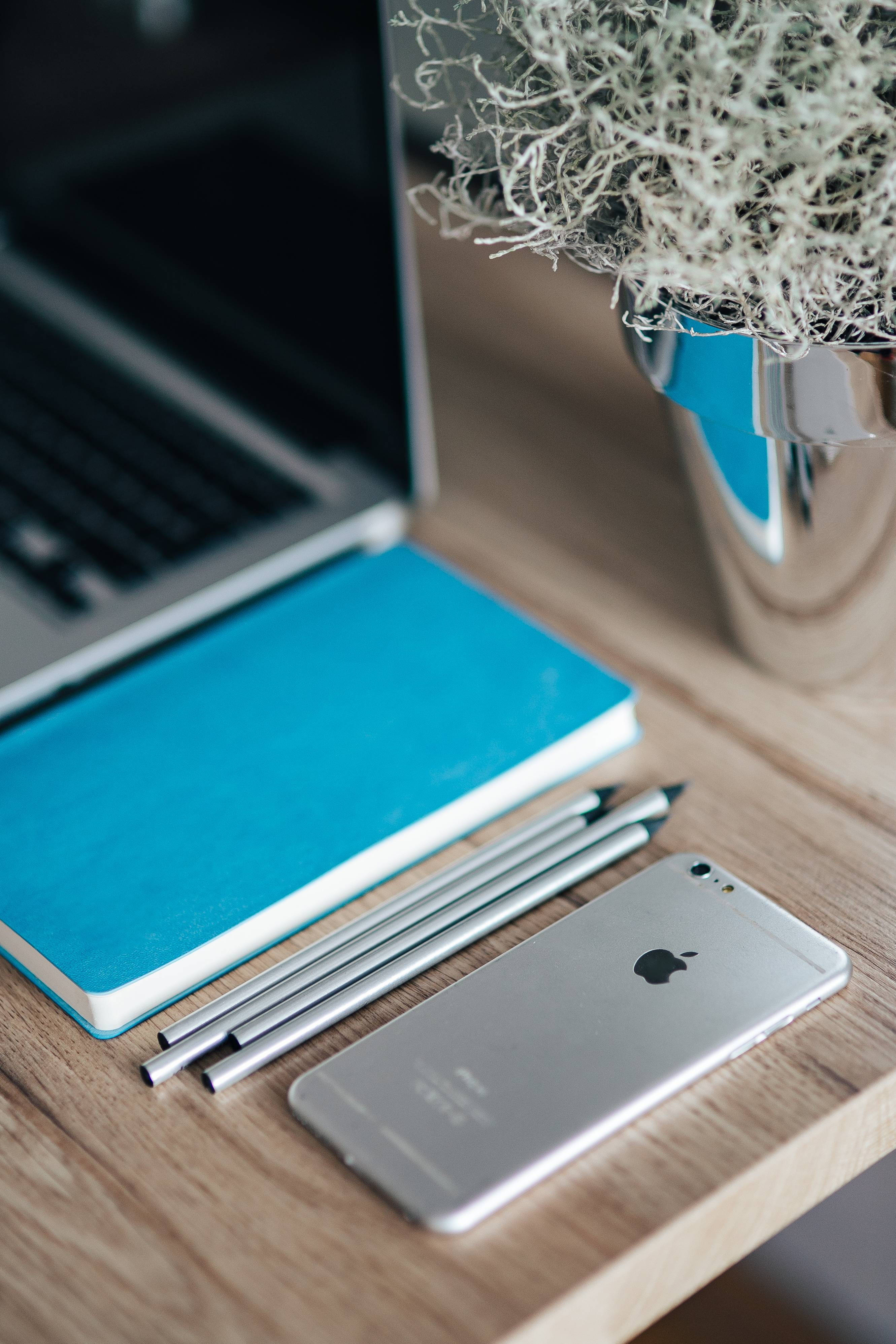 MacBook 動態圖片 桌布 壁紙 — 一張簡約木桌上放著銀色筆記型電腦、藍色筆記本、鉛筆和電話，旁邊有一個銀色花瓶，裡面有白色苔蘚。