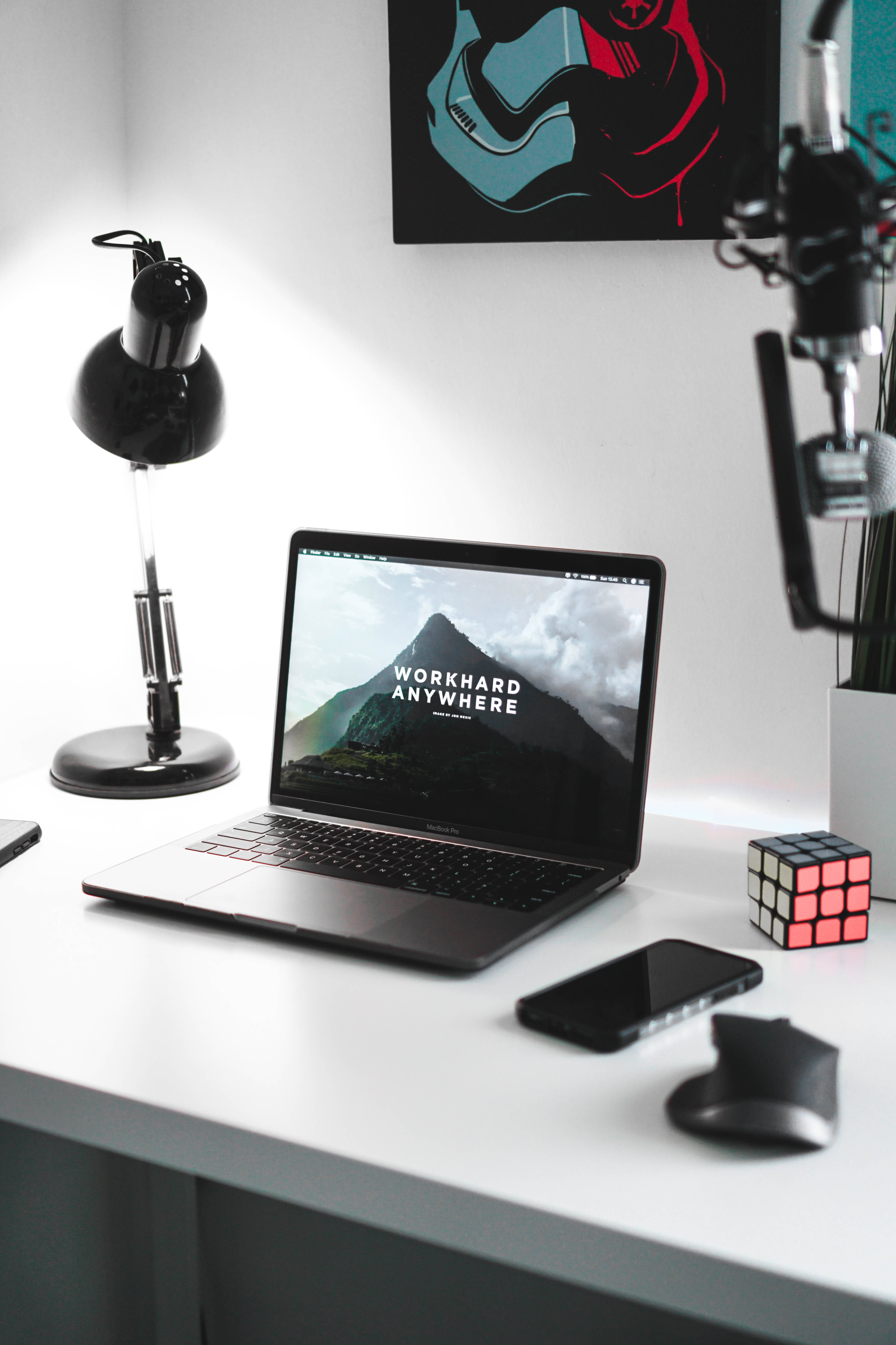 MacBook 動態圖片 桌布 壁紙 — 一張簡約風格的白色桌子上放著一台筆記型電腦，顯示著一幅山景桌布，上面有 workhard anywhere 字樣，旁邊有黑色檯燈、電話、滑鼠和魔術方塊。