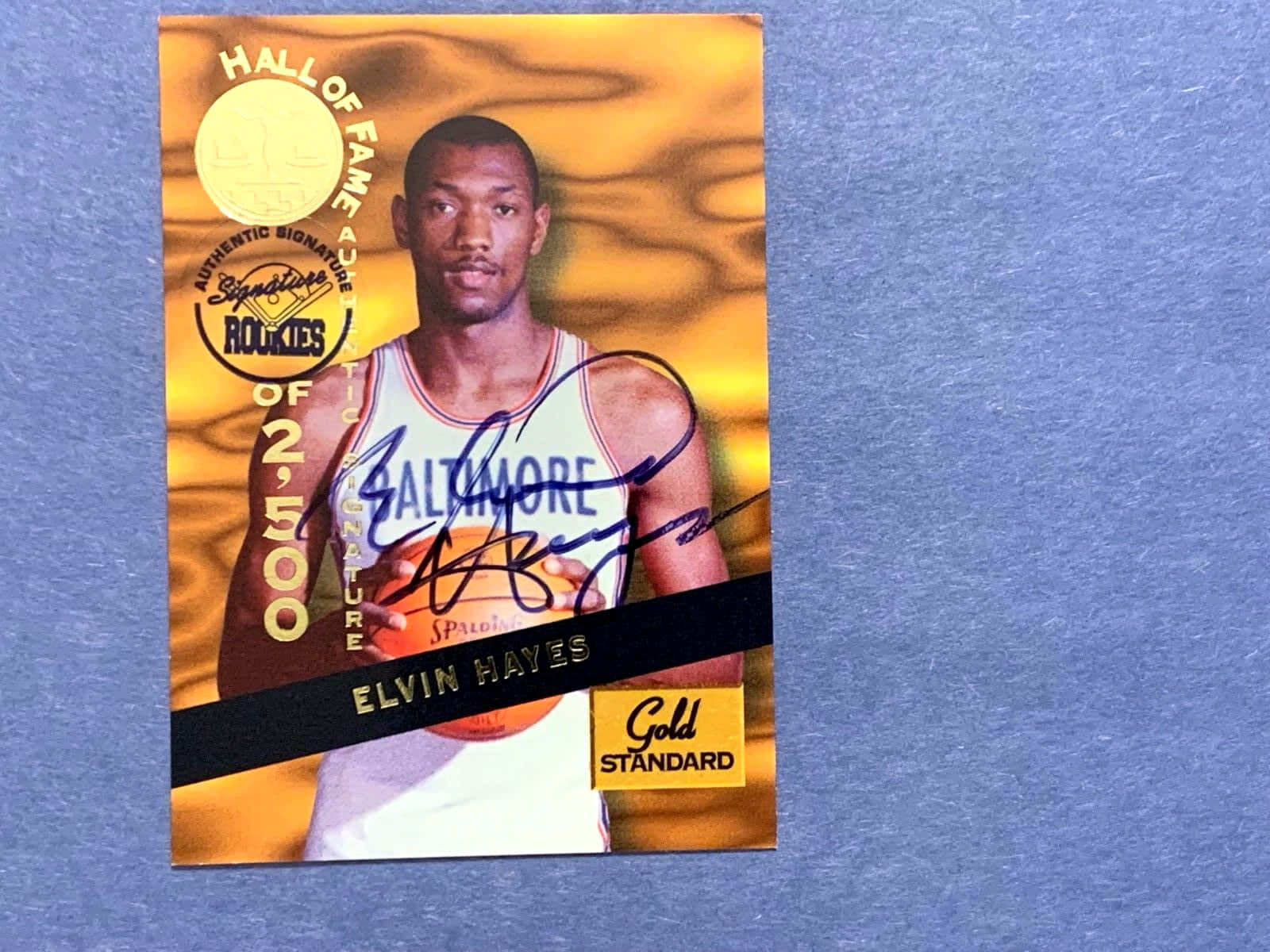 籃球 桌布 壁紙 — 一張簽名的 Elvin Hayes 名人堂新秀卡，金色箔紙背景，放在有紋理的藍色表面上。