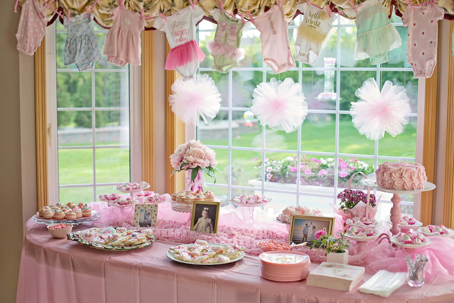 Baby Shower 桌布 壁紙 — 一張粉彩粉紅色嬰兒派對甜品桌，裝飾有杯子蛋糕、蛋糕和芭蕾舞裙裝飾，位於可欣賞綠色花園景觀的窗戶下方。