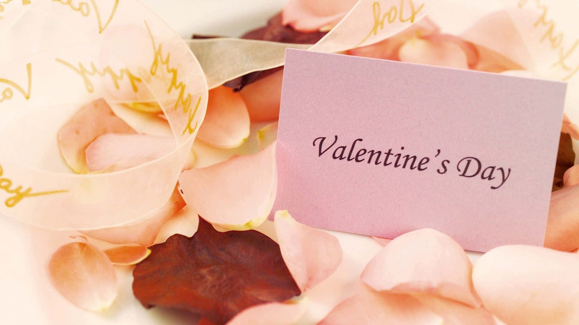Valentines 桌布 壁紙 — 一張粉紅色情人節卡片，藏在柔軟的玫瑰花瓣和金色緞帶之間，帶有手寫字體。