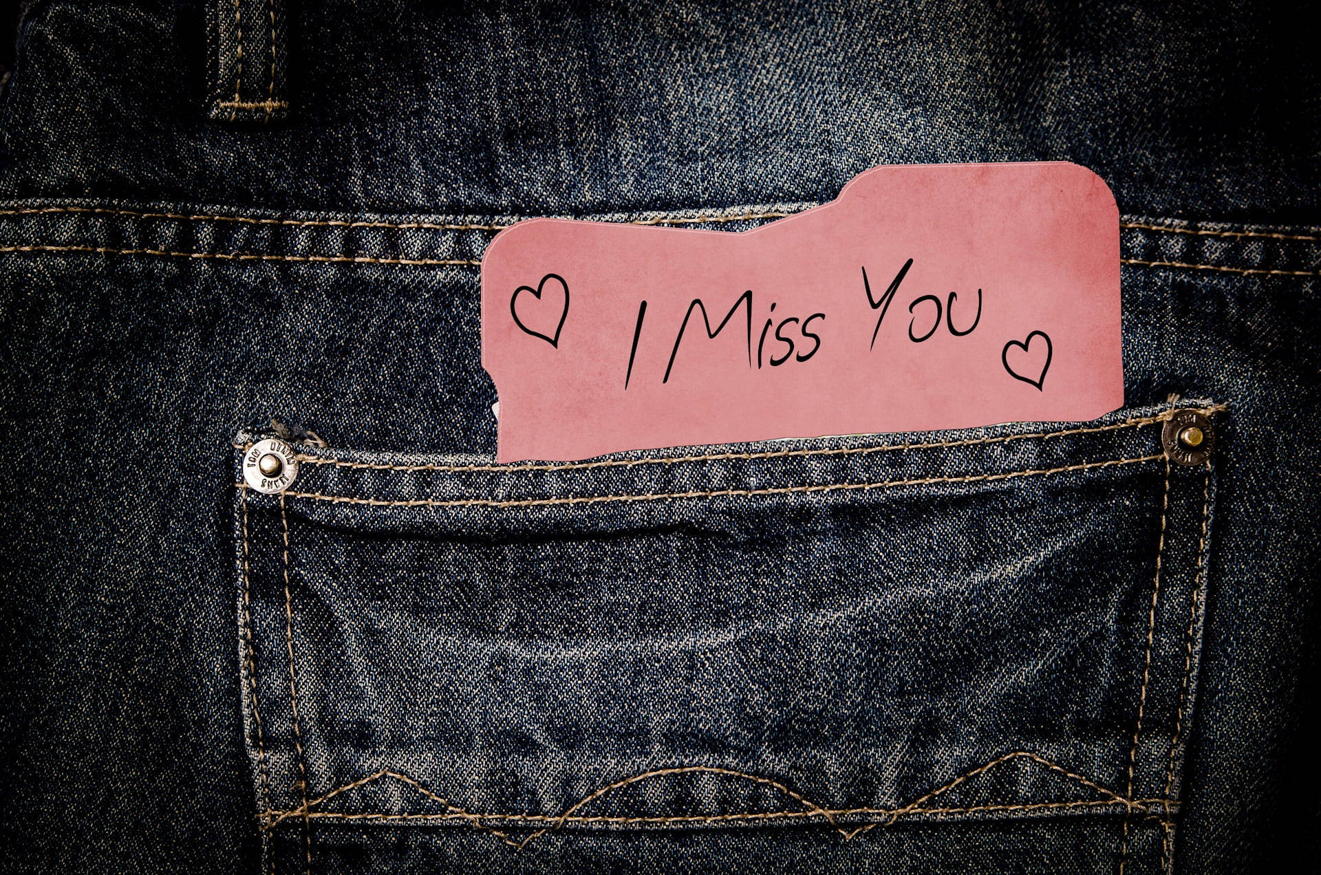 Missing 桌布 壁紙 — 一張粉紅色手寫便條，上面寫著「I Miss You」並帶有心形圖案，塞在深色牛仔褲的後袋裡。