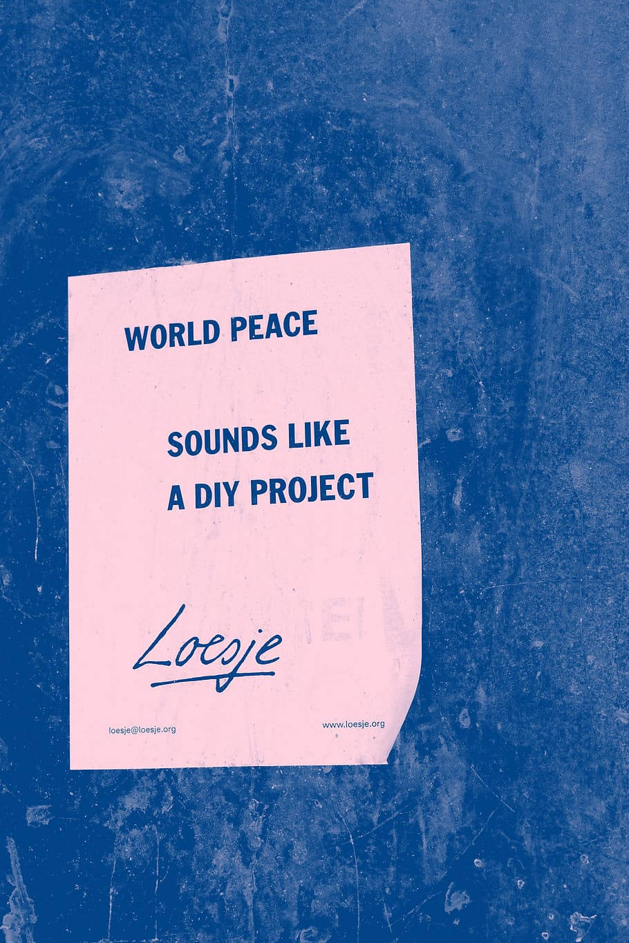 iPhone 桌布 壁紙 — 一張粉紅色紙，上面有藍色文字寫著「WORLD PEACE SOUNDS LIKE A DIY PROJECT」，由Loeije創作，用膠帶貼在紋理藍色牆上。