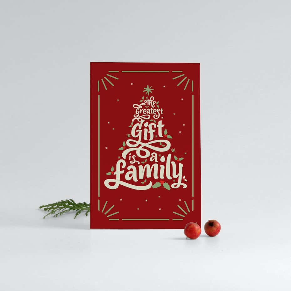 Christmas Card 桌布 壁紙 — 一張紅色節日賀卡，上面有白色草書文字寫著「The Greatest Gift is a Family」，周圍環繞著冬青和金飾，旁邊有松枝和紅色漿果放在白色表面上。
