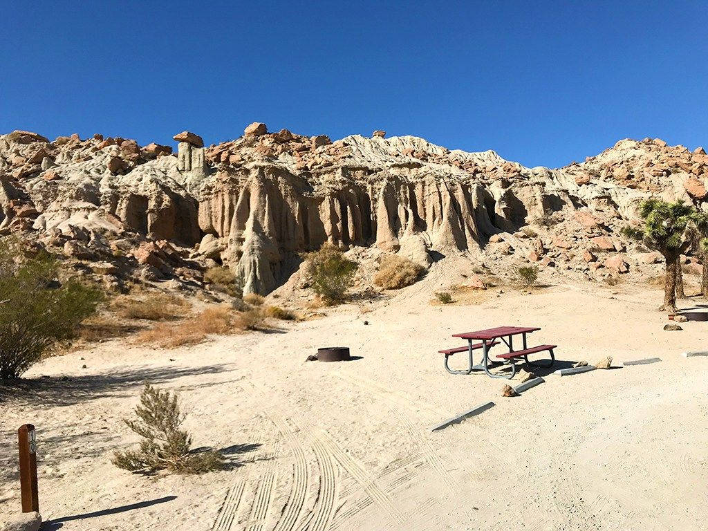 rocky 桌布 壁紙 — 一張紅色野餐桌放在陽光明媚的沙漠營地中，周圍有被侵蝕的岩石 formations 和一棵孤獨的棕櫚樹，在清澈的藍天下。
