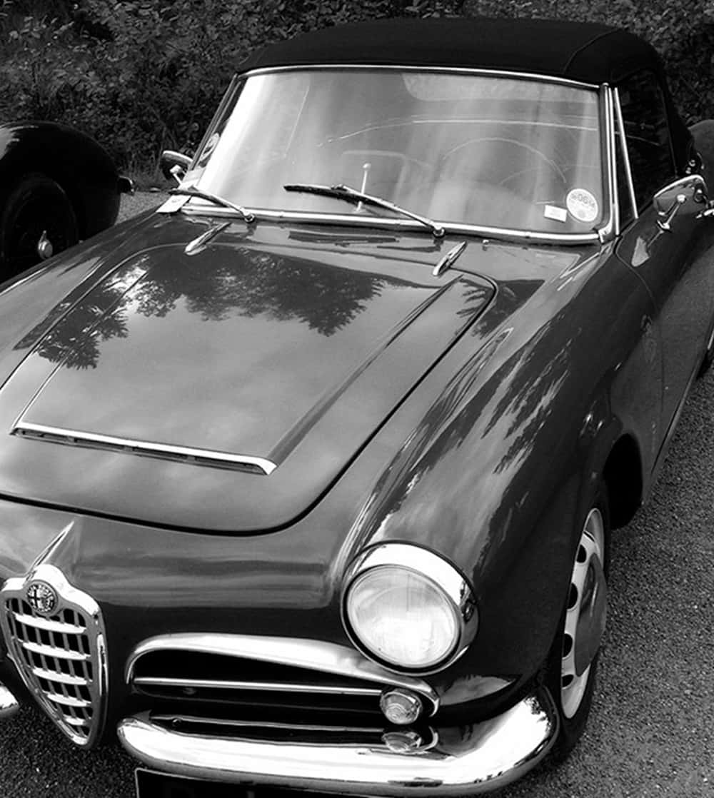 Car Hood 桌布 壁紙 — 一張經典黑白照片，一輛復古愛快羅密歐敞篷車停放在戶外，樹木倒映在其光亮的引擎蓋上。