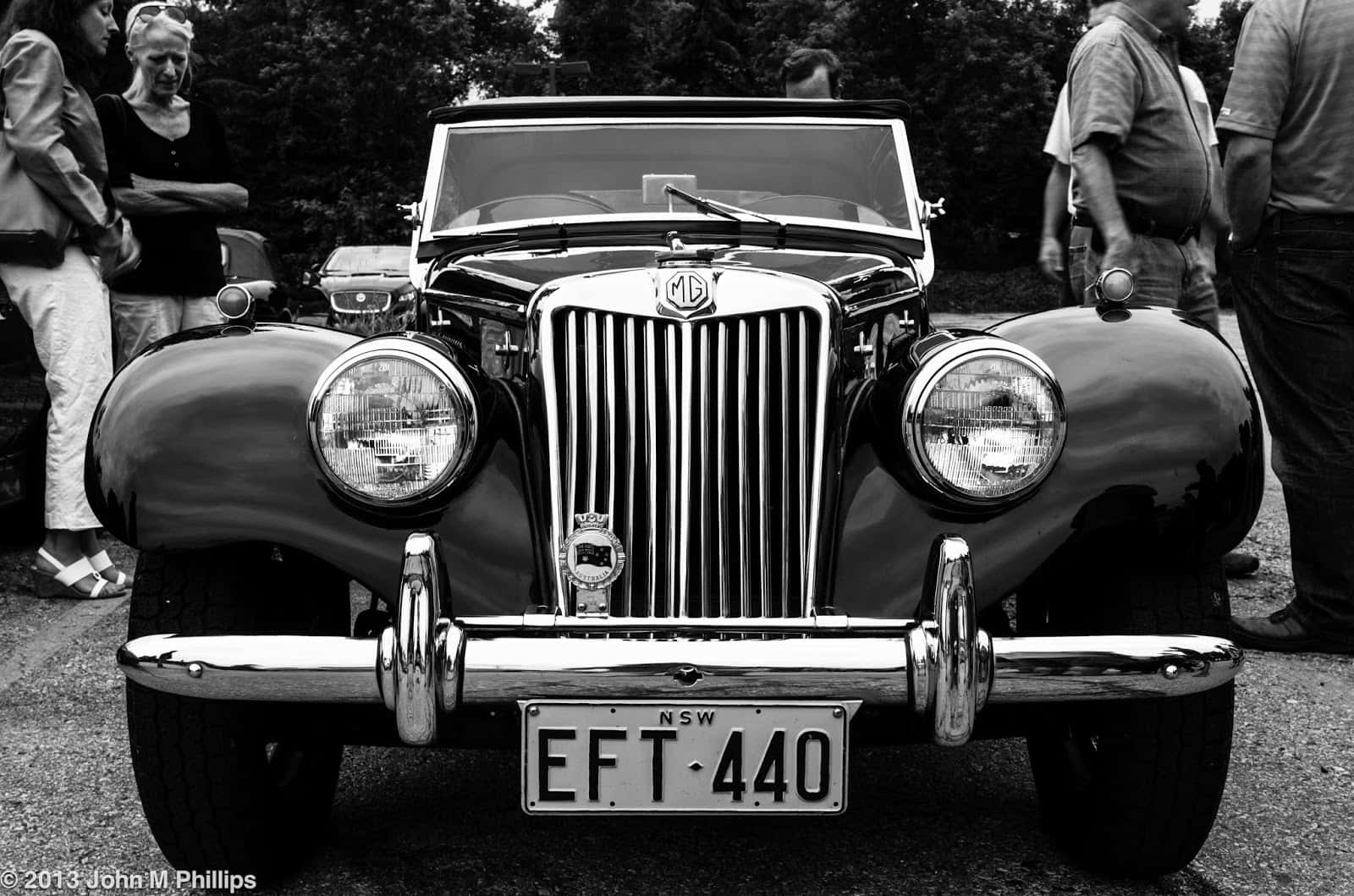 Black And White Vintage Car 桌布 壁紙 — 一張經典黑白照片，展示一輛配備鍍鉻格柵和圓形頭燈的復古MG敞篷車，在戶外聚會中被圍觀者包圍。