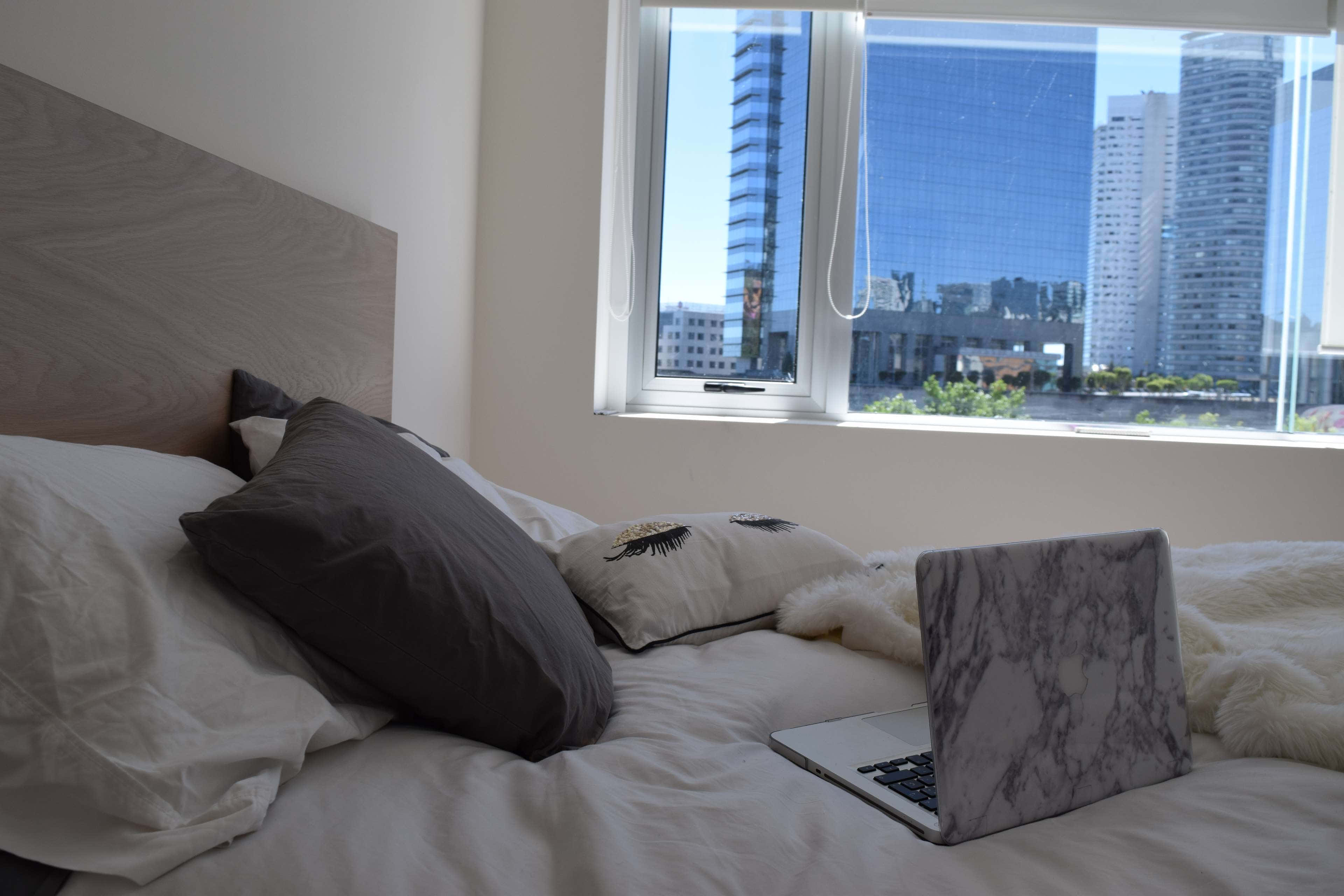 Hotel Room 桌布 壁紙 — 一張舒適的床，配有白色床單和灰色枕頭，面向一扇窗戶，窗外是明亮的天際線，高聳的玻璃建築林立。