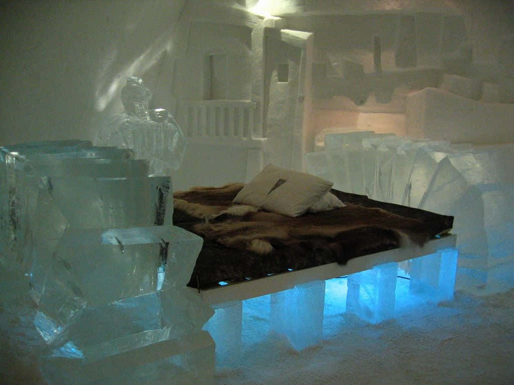 Ice Hotel 桌布 壁紙 — 一張舒適的床，鋪有毛皮和枕頭，放置在發光的藍色冰塊上，位於雕刻精美的冰室內。