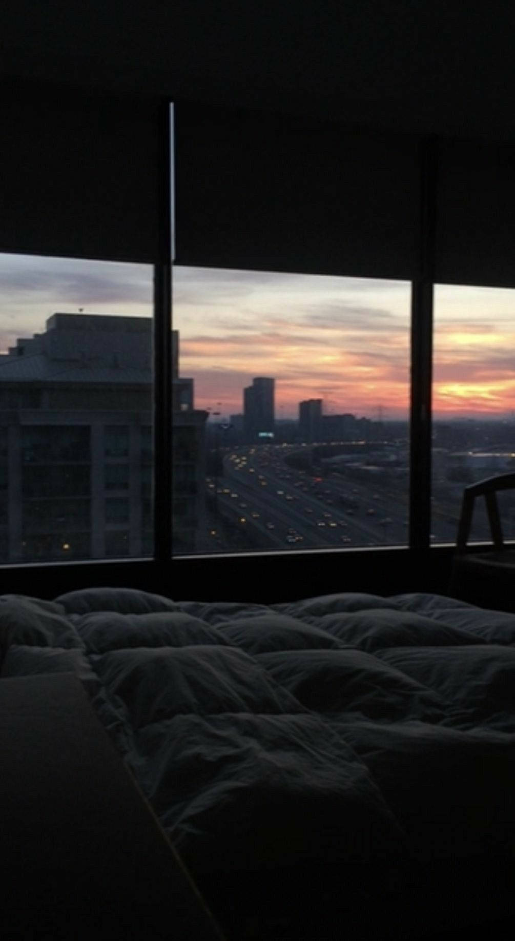 room 桌布 壁紙 — 一張舒適的床俯瞰著日落時分的城市景觀，橙色和粉紅色的雲彩條紋在發光的高速公路車流上方。