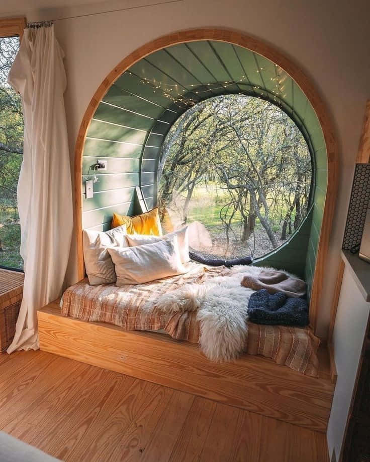cabin 桌布 壁紙 — 一張舒適的木床嵌在圓形窗框內，俯瞰陽光普照的森林，配有燈串和柔軟的毯子。