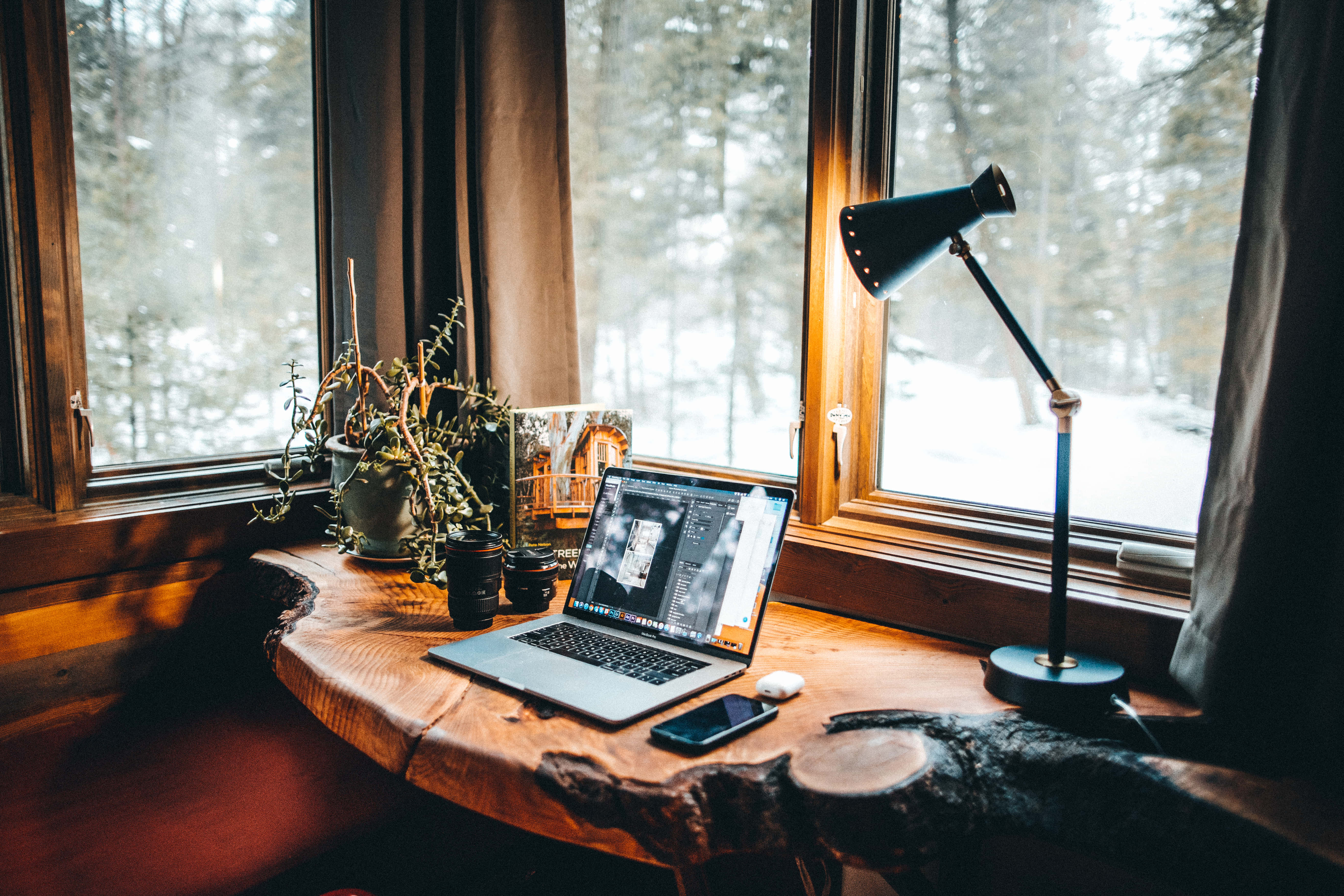 Cozy Winter Desktop 桌布 壁紙 — 一張舒適的木桌放在雪窗前，桌上放著筆記型電腦、燈、植物和手機，沐浴在溫暖的金色光芒中。