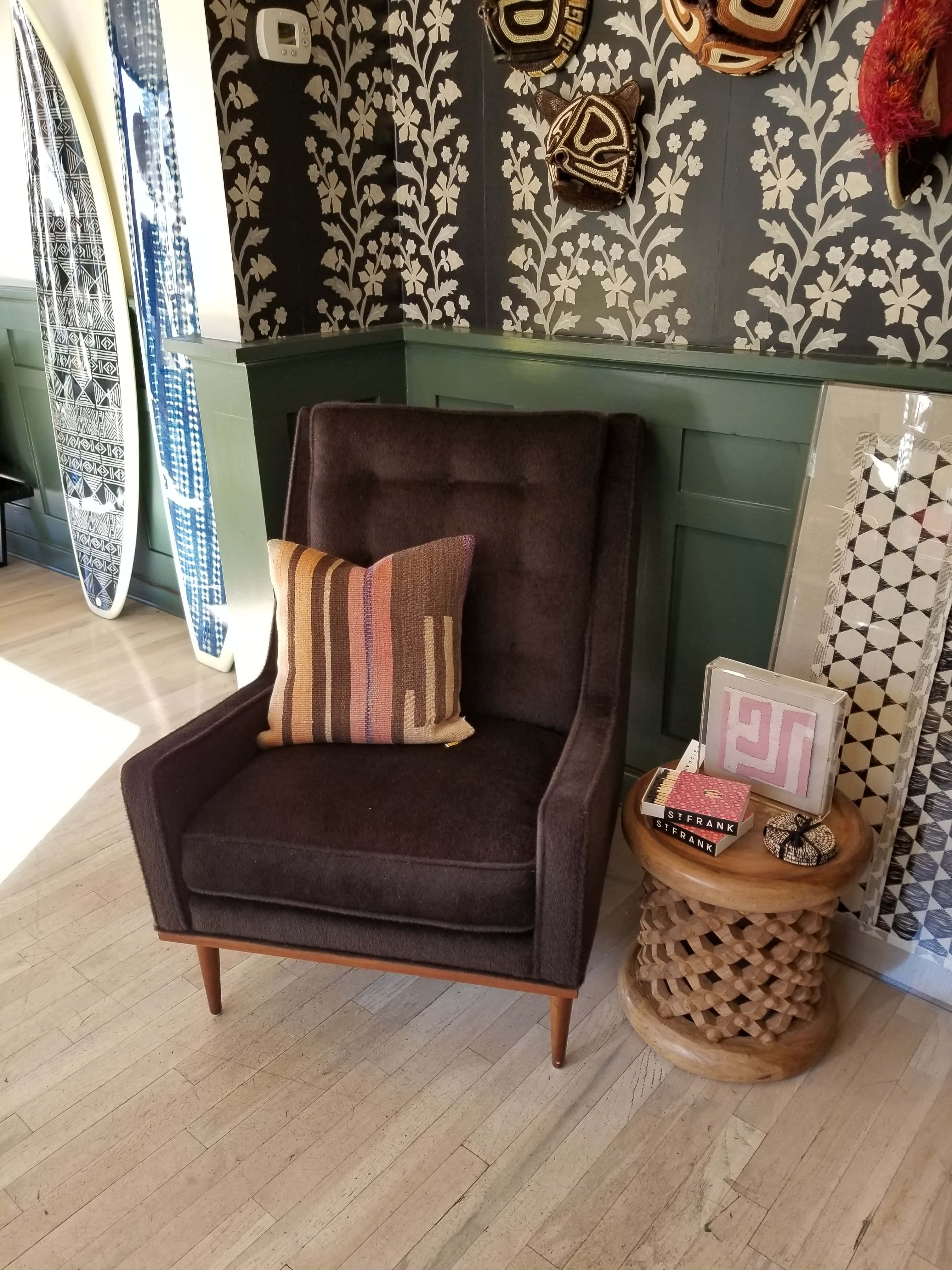 Interior Design 桌布 壁紙 — 一張舒適的棕色扶手椅配以條紋靠墊，旁邊放著一張編織邊桌，房間內有圖案壁紙和靠在牆上的衝浪板。