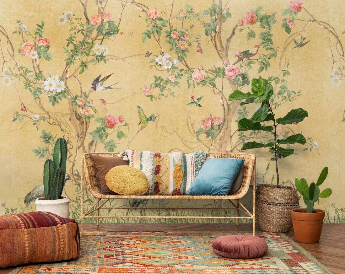 Chinoiserie 桌布 壁紙 — 一張藤編沙發配以色彩繽紛的靠墊，置於描繪鳥兒和開花樹木的暖色調花卉壁紙前。