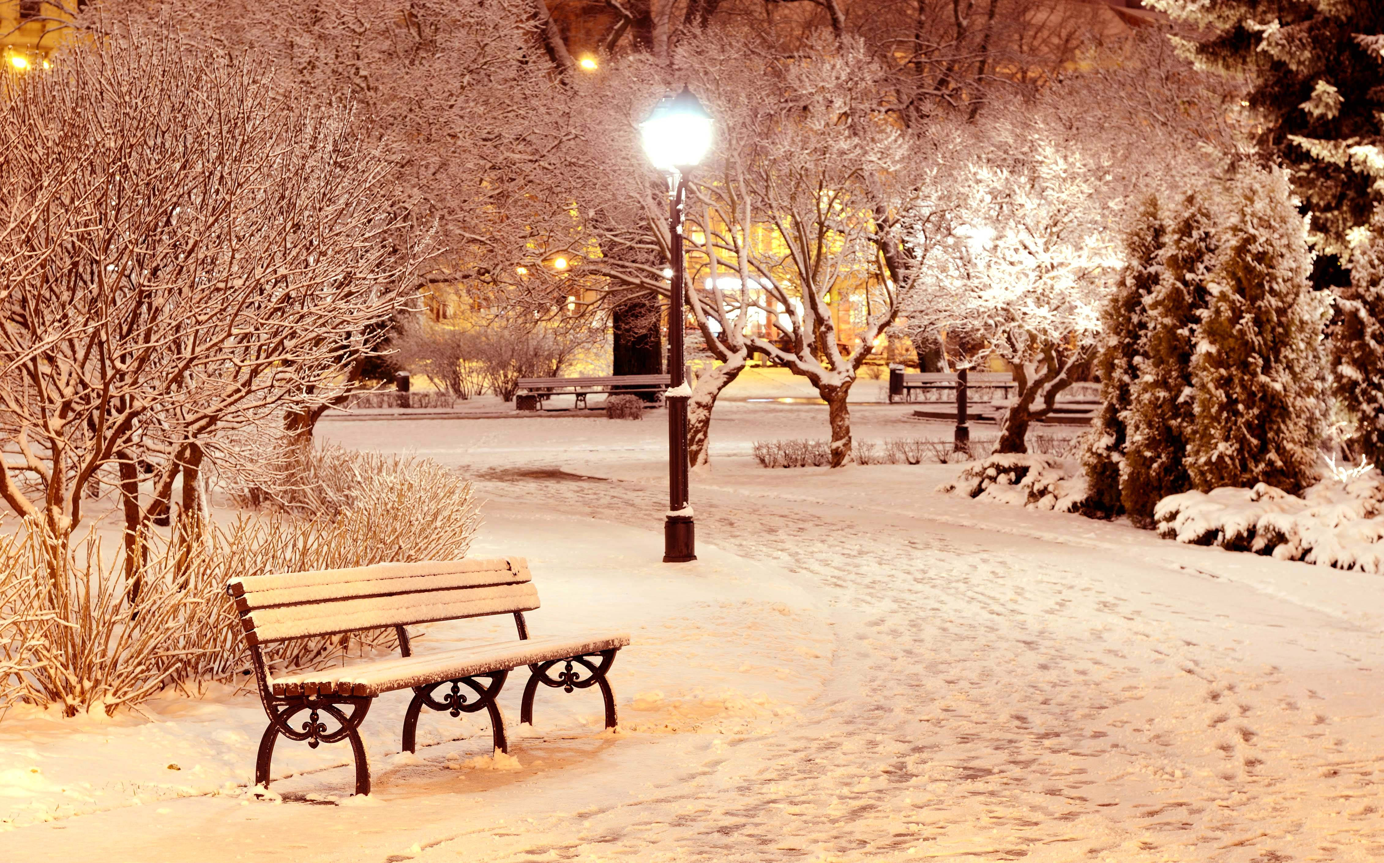 冬季 桌布 壁紙 — 一張被雪覆蓋的公園長椅在夜晚的路燈下溫暖地發光，周圍有結霜的樹木和柔軟白雪上的腳印。