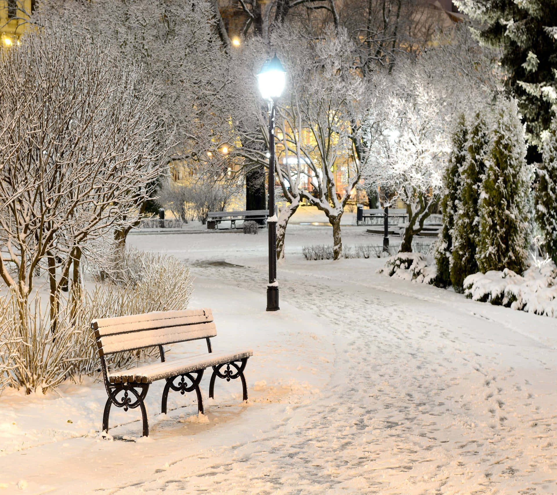 Cozy Winter Desktop 桌布 壁紙 — 一張被雪覆蓋的公園長椅在夜晚的路燈下發光，周圍有結霜的樹木和柔軟白雪上的腳印。