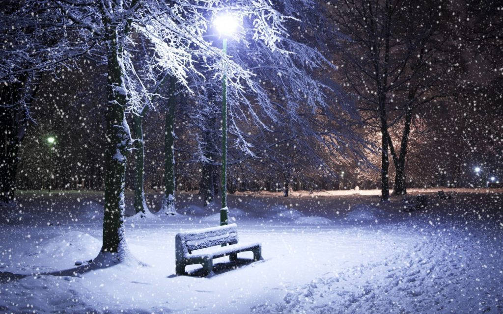 冬季桌面 桌布 壁紙 — 一張被雪覆蓋的公園長椅在街燈下發光，夜間雪花輕輕飄過光禿的冬季樹木。