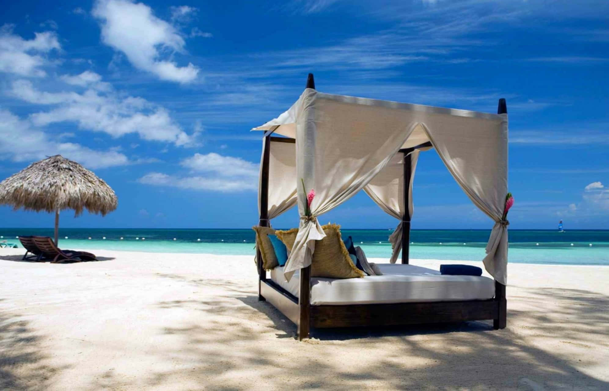 Beach Daybed 桌布 壁紙 — 一張豪華的華蓋床放置在純淨的白色沙灘上，上方是充滿活力的藍天，點綴著蓬鬆的雲朵和碧綠的海浪。