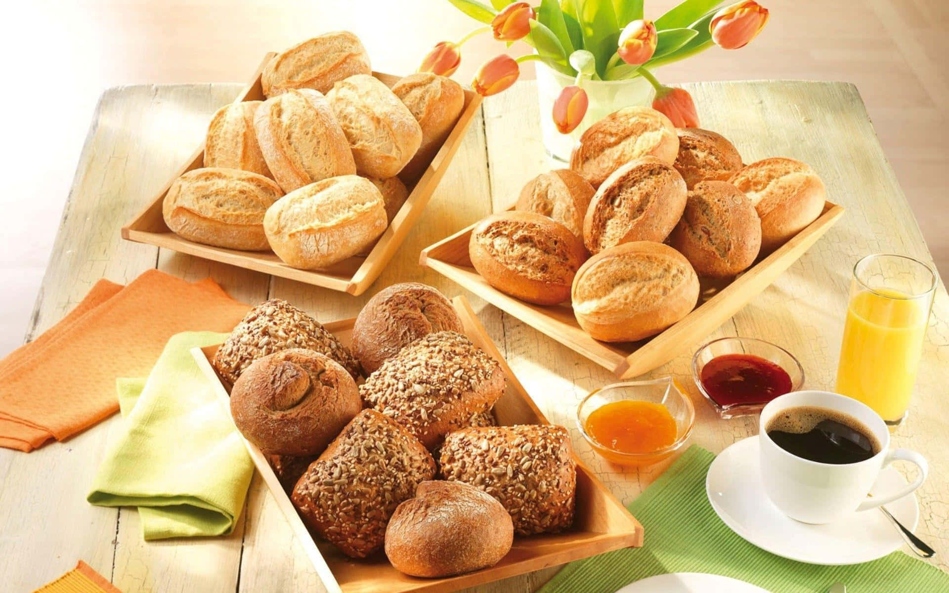 Bakery 桌布 壁紙 — 一張質樸的木桌上，在溫暖的晨光下展示著三盤各式新鮮麵包，搭配橙色鬱金香和早餐食品。