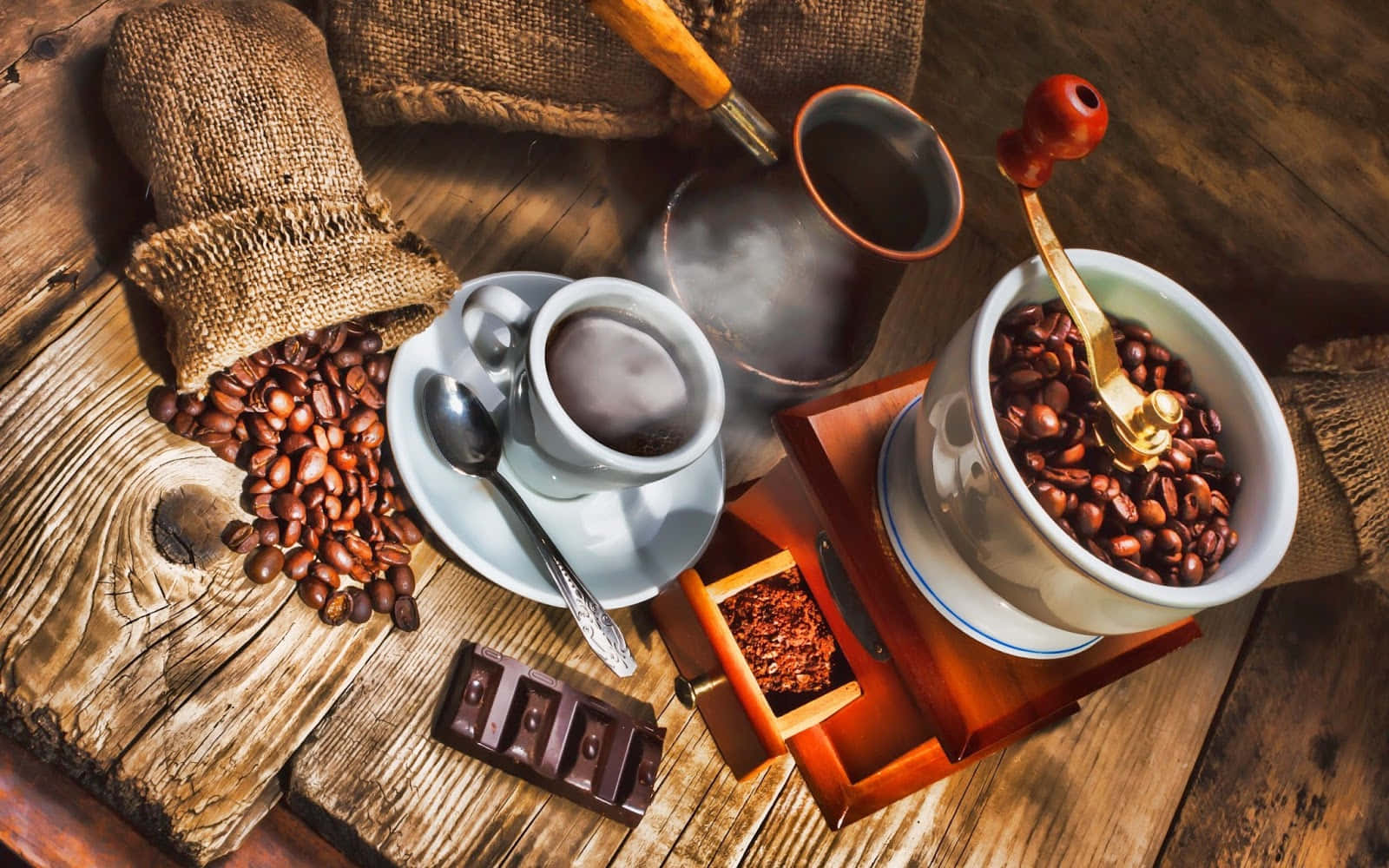 Coffee Aesthetic 桌布 壁紙 — 一張質樸的木桌上展示了一杯冒煙的咖啡、麻布袋中的咖啡豆、巧克力條和手動研磨機，沐浴在溫暖的金色光芒中。