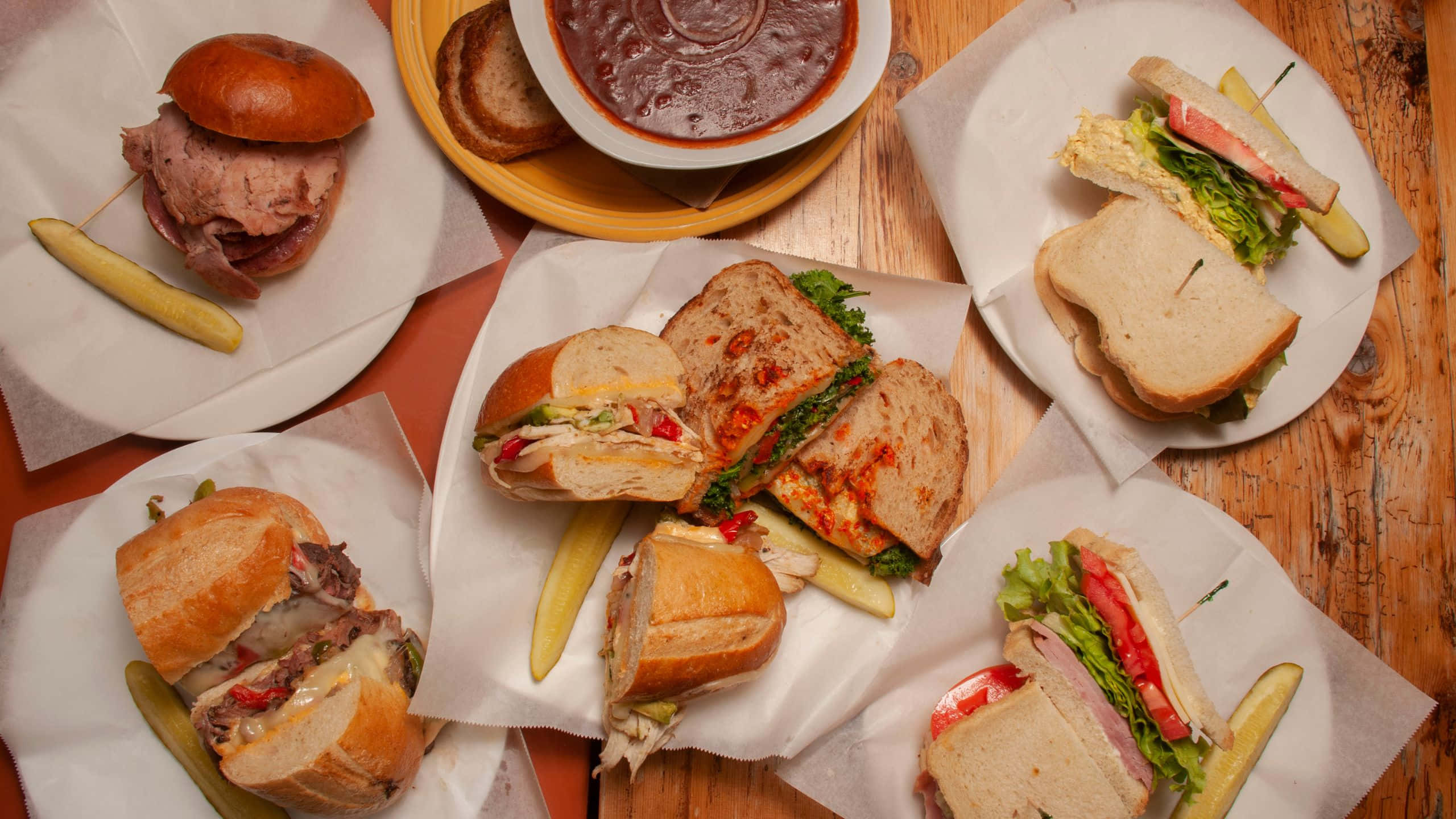 Delicious Food 桌布 壁紙 — 一張質樸的木桌上展示了一系列新鮮製作的三明治和一碗濃郁的醬汁，每份都配有醃黃瓜，放在烘焙紙上。