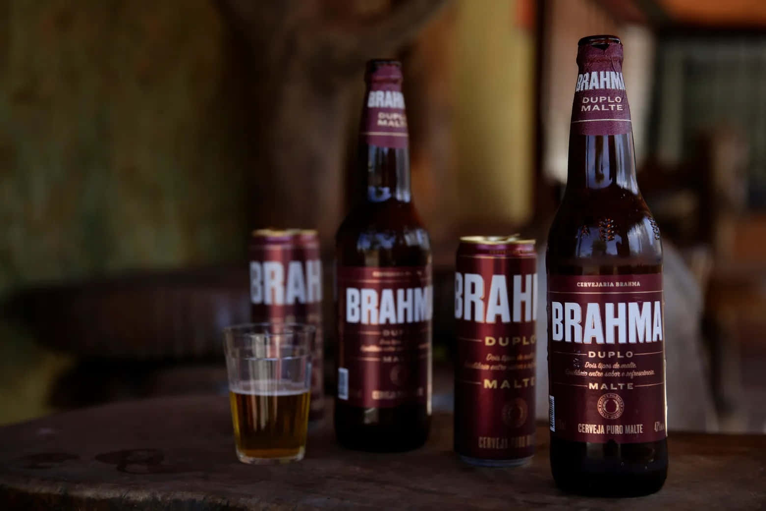 alcohol 桌布 壁紙 — 一張質樸的木桌上展示著 Brahma 啤酒瓶、罐和一杯琥珀色啤酒，在溫暖的環境光線中。
