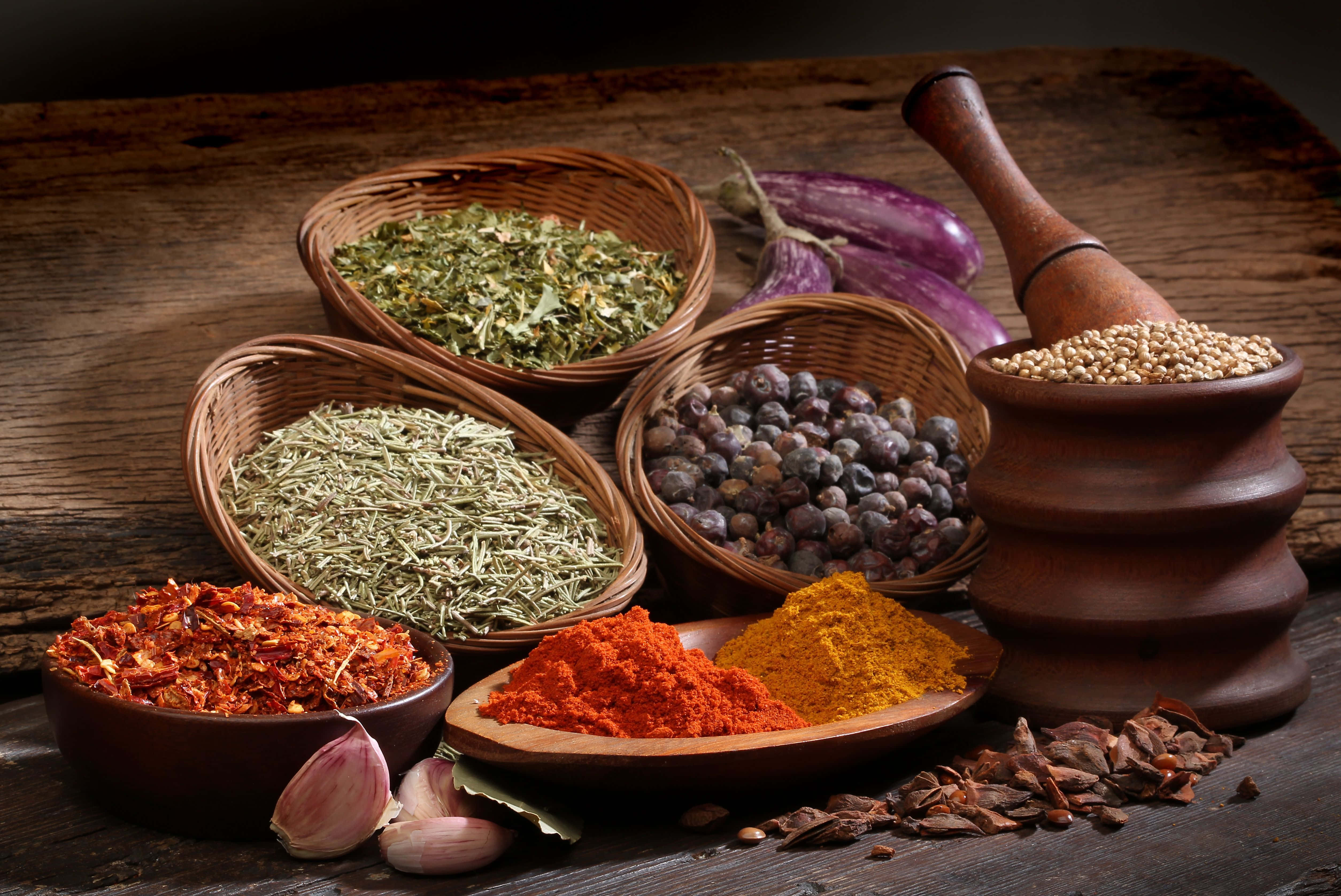 Spices 桌布 壁紙 — 一張質樸的木桌上展示著編織籃子和碗中的鮮豔香料，還有大蒜、茄子和一個散落種子的研缽。