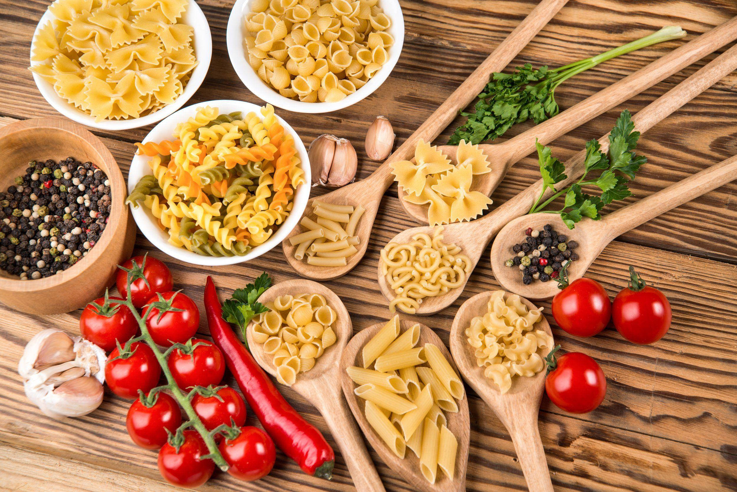 Food Photography 桌布 壁紙 — 一張質樸的木桌上展示著色彩繽紛的義大利麵、新鮮番茄、大蒜和香草，擺放在木勺和碗中。